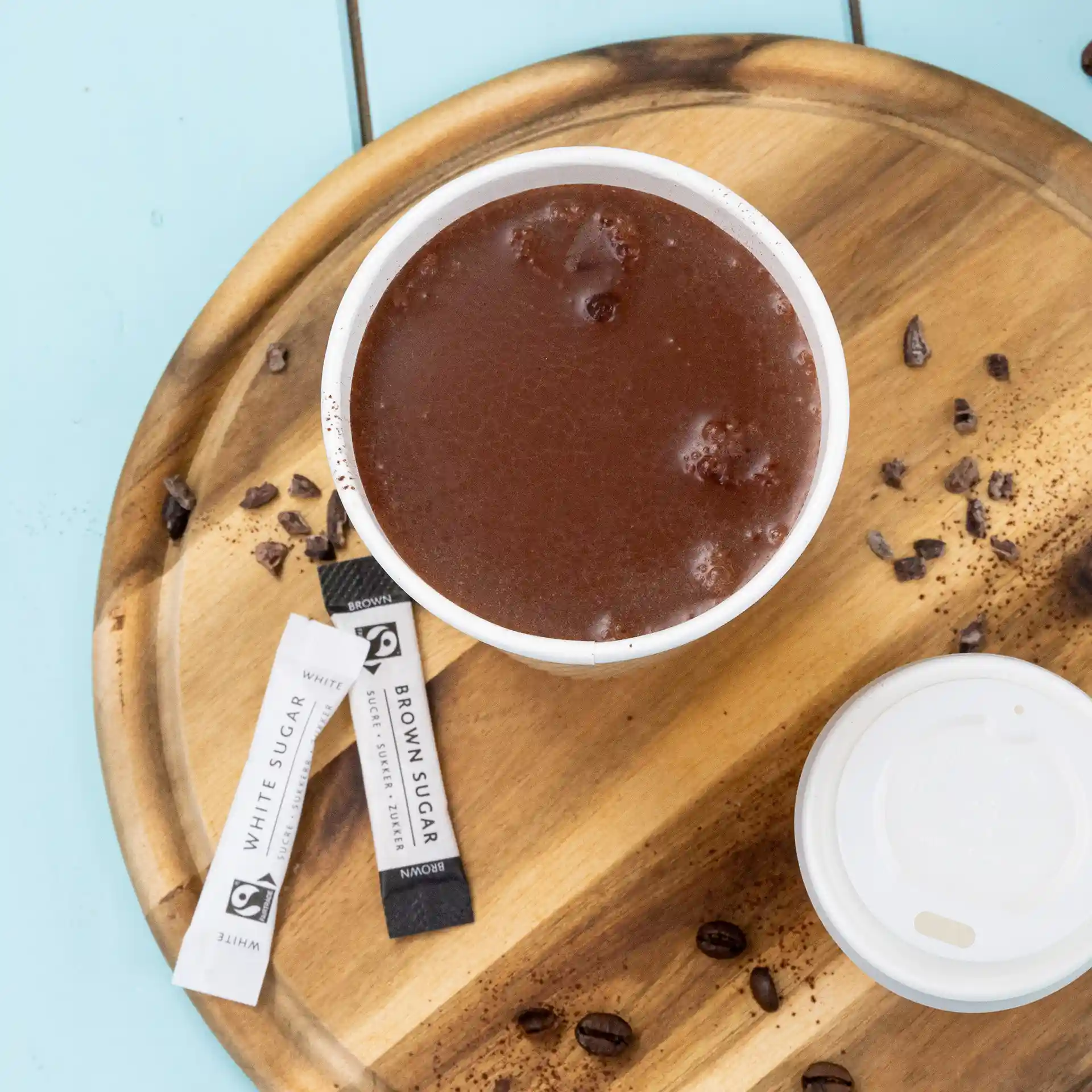 Beker warme chocolademelk op een houten dienblad met suikerzakjes en verspreide koffiebonen.