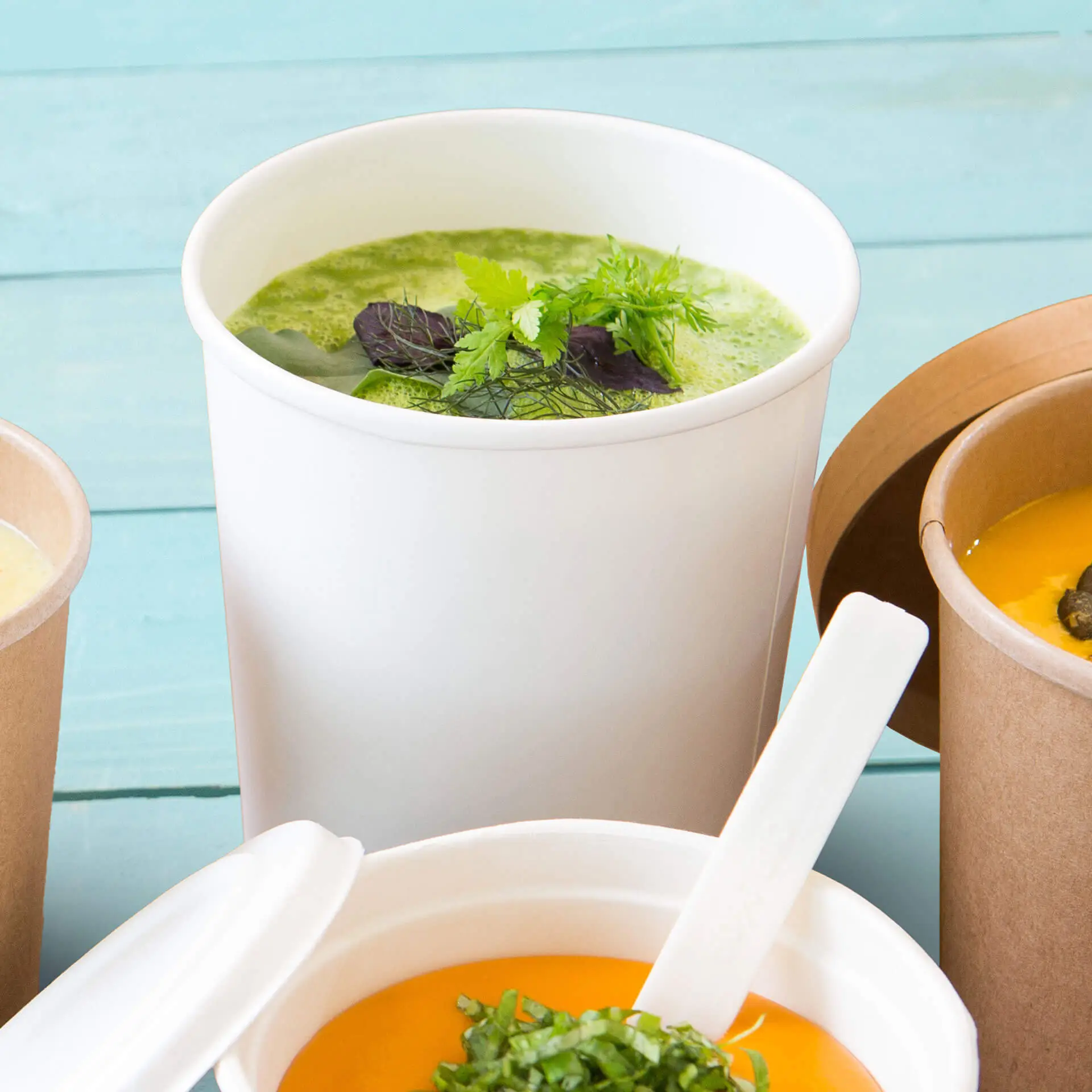 Witte biologisch afbreekbare soepbeker met groene soep en kruiden, geplaatst met andere soepcontainers op een tealte achtergrond.