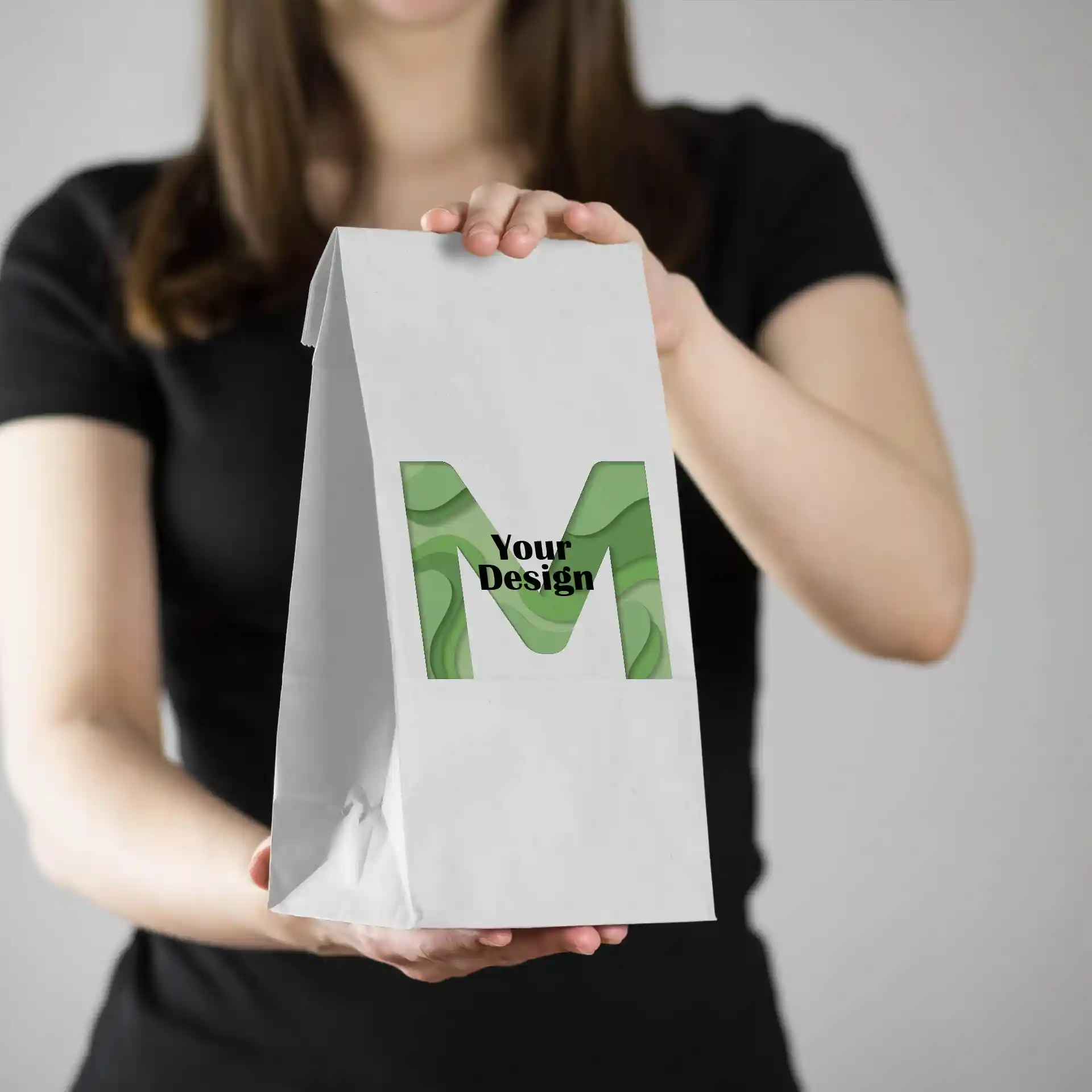 Witte papieren tas vastgehouden door een persoon, met een groen ontwerp met de letter 'M' en de tekst 'Jouw Ontwerp'.