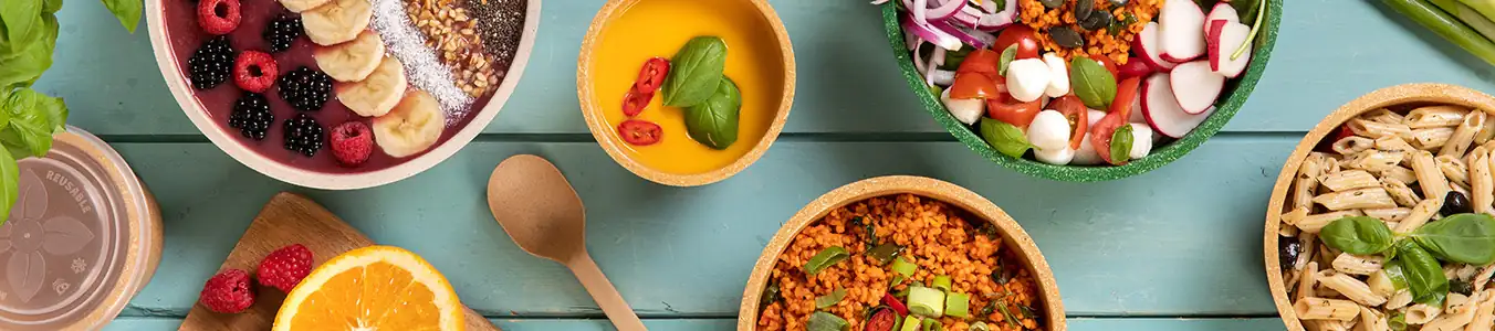 Kleurvolle kommen gevuld met diverse voedingsmiddelen, waaronder fruit, pasta, salade en granen, op een houten tafel gerangschikt.