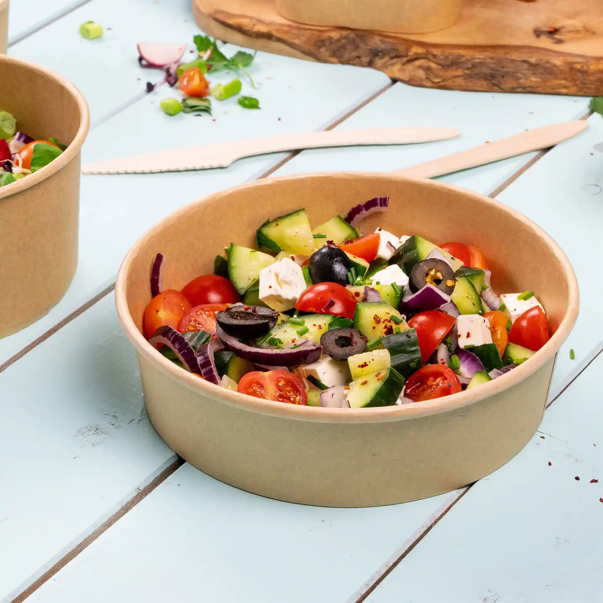 Ronde bruine kartonnen kom gevuld met salade, inclusief tomaten, komkommers, olijven en feta, op een blauwe ondergrond.
