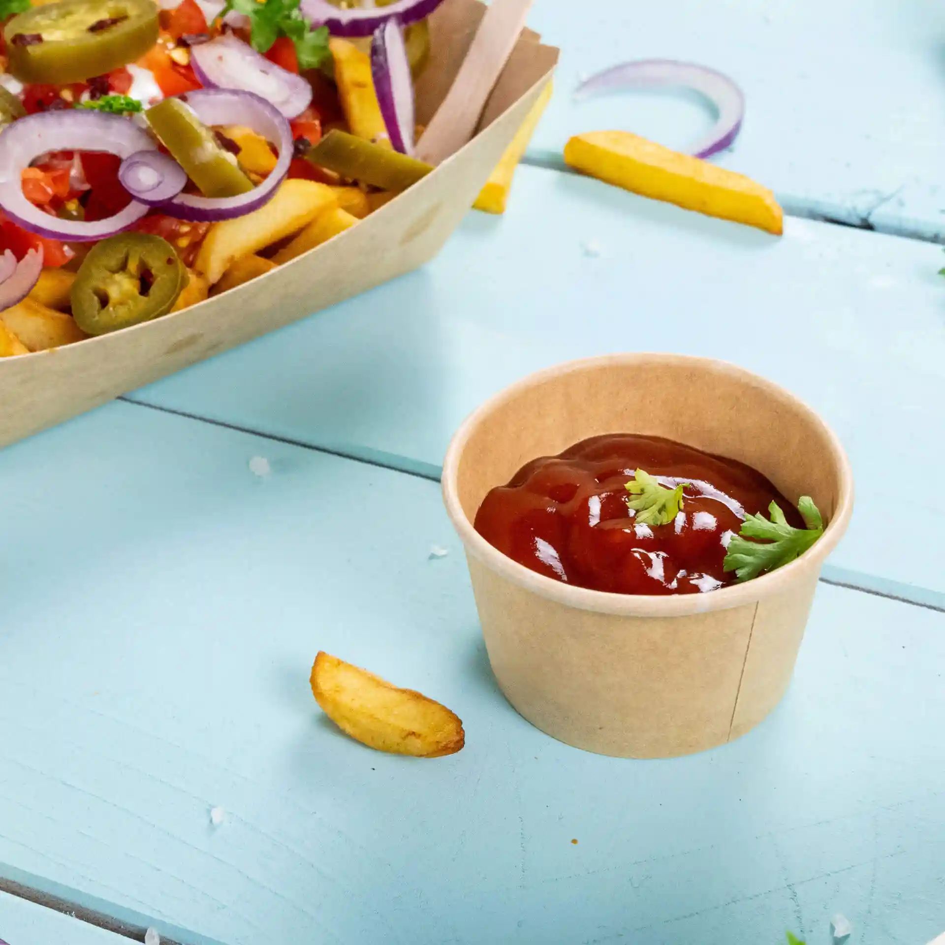 Bruine papieren portiebeker met ketchup en een garnering van koriander, op een lichtblauwe ondergrond met frietjes erbij.