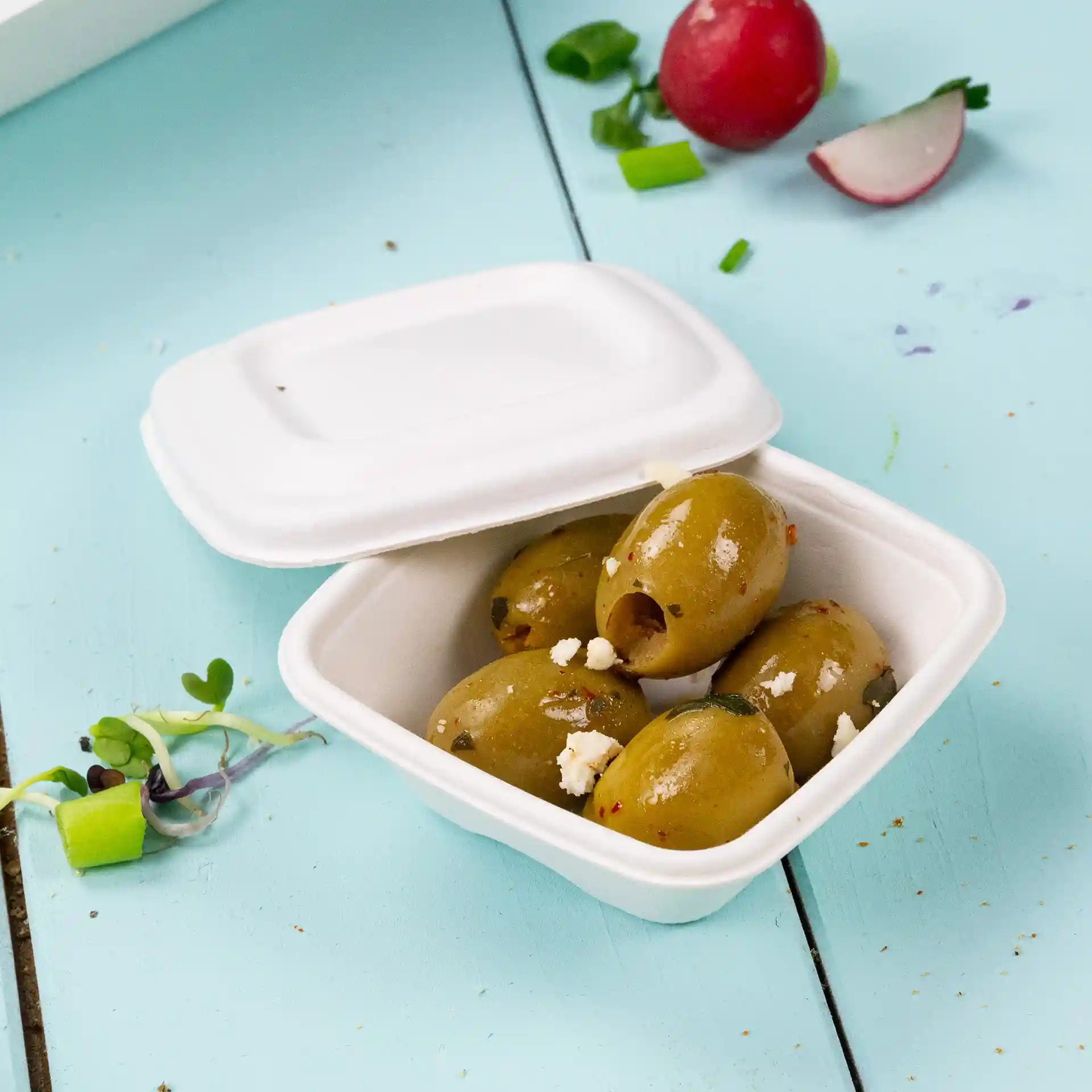 Container met verschillende groene olijven erin, geplaatst op een lichtblauwe ondergrond, met een gedeeltelijk gesloten wit deksel.