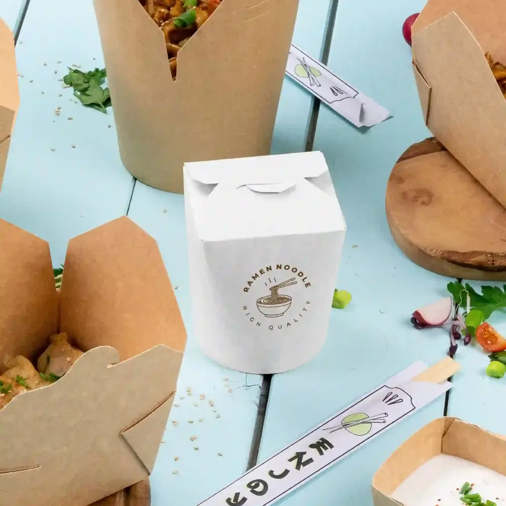 Witte afhaaldoos met het label "Ramen Noodles van hoge kwaliteit" naast bruine afhaaldozen op een blauwe ondergrond.