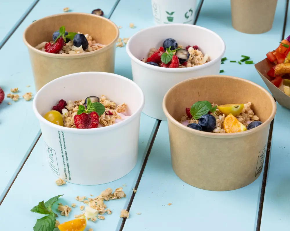 Vier voedselcontainers gevuld met granola en vers fruit, waaronder bessen en citrus, op een blauwe houten ondergrond.