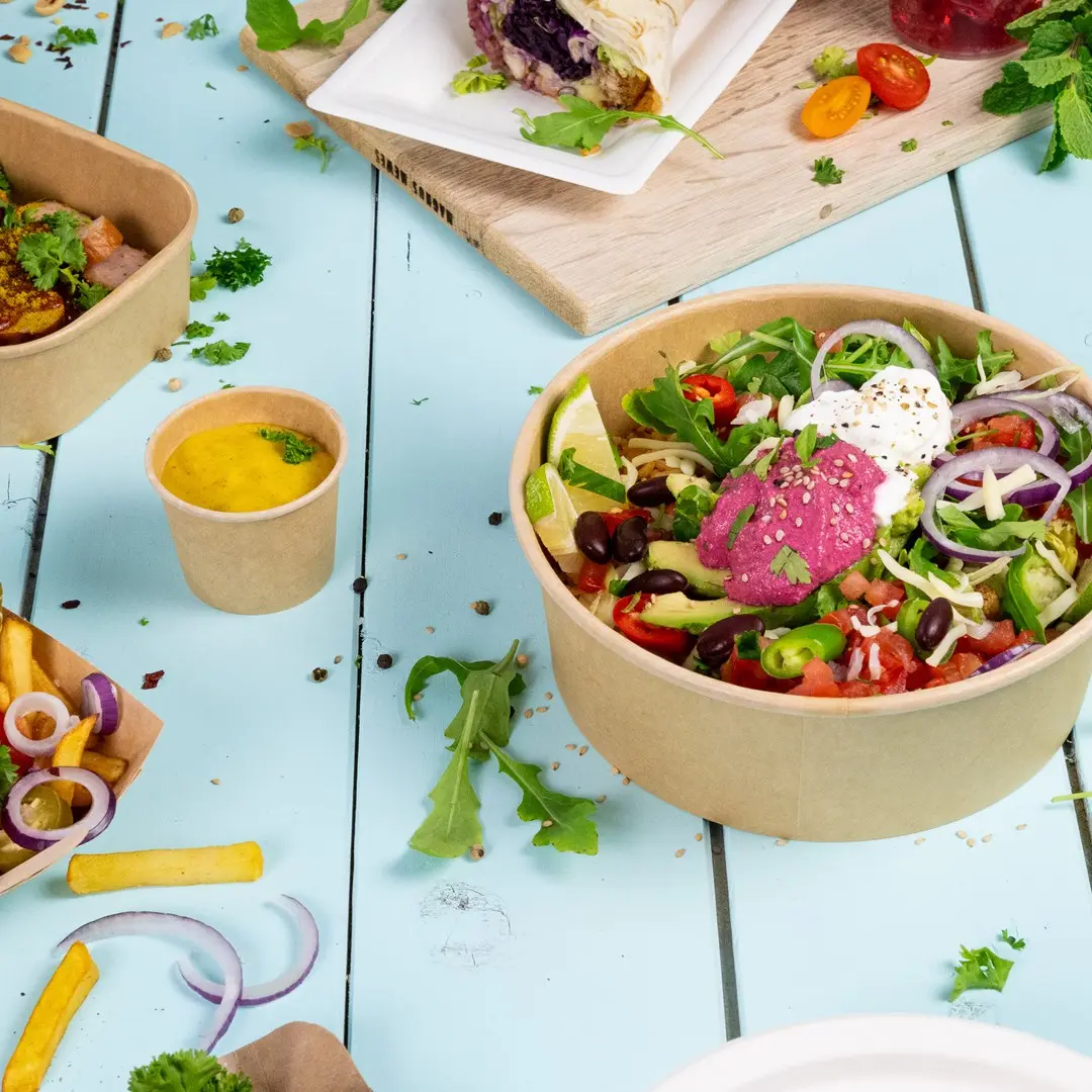 Kom met salade met gemengde groenten, limoen en sauzen, omringd door kleinere containers met saus, friet en kruiden op een blauwe ondergrond.