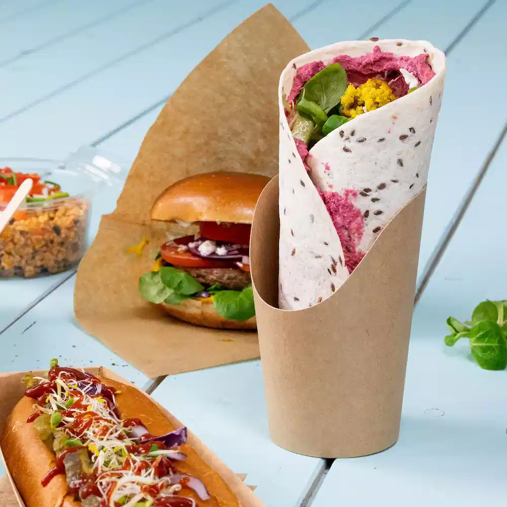 Wrap gevuld met groenten en spread, geserveerd in een bruine papieren houder, met een burger en hotdog op de achtergrond.