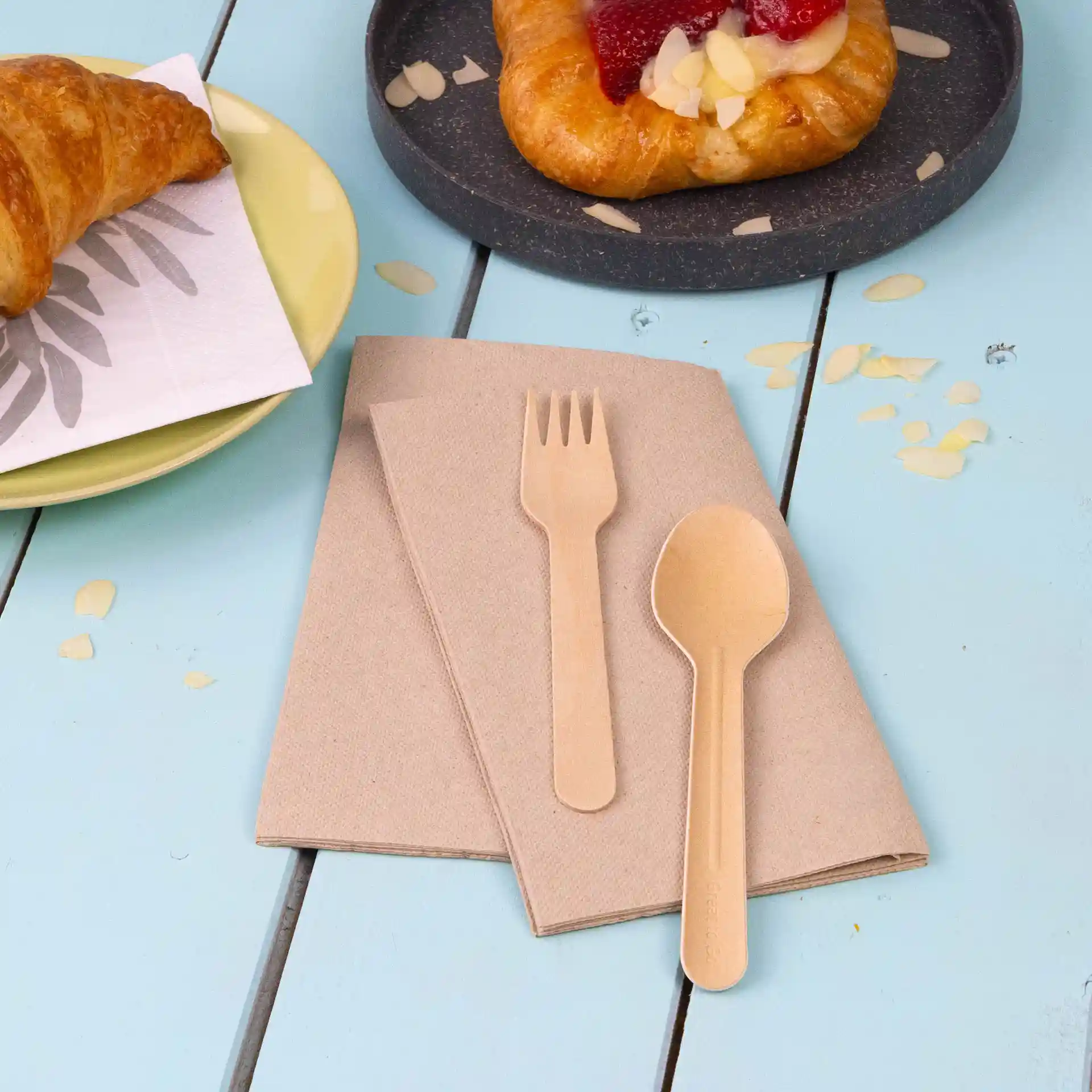 Twee ongebleekte gerecycleerde papieren servetten met een houten vork en lepel op een blauwe tafel.