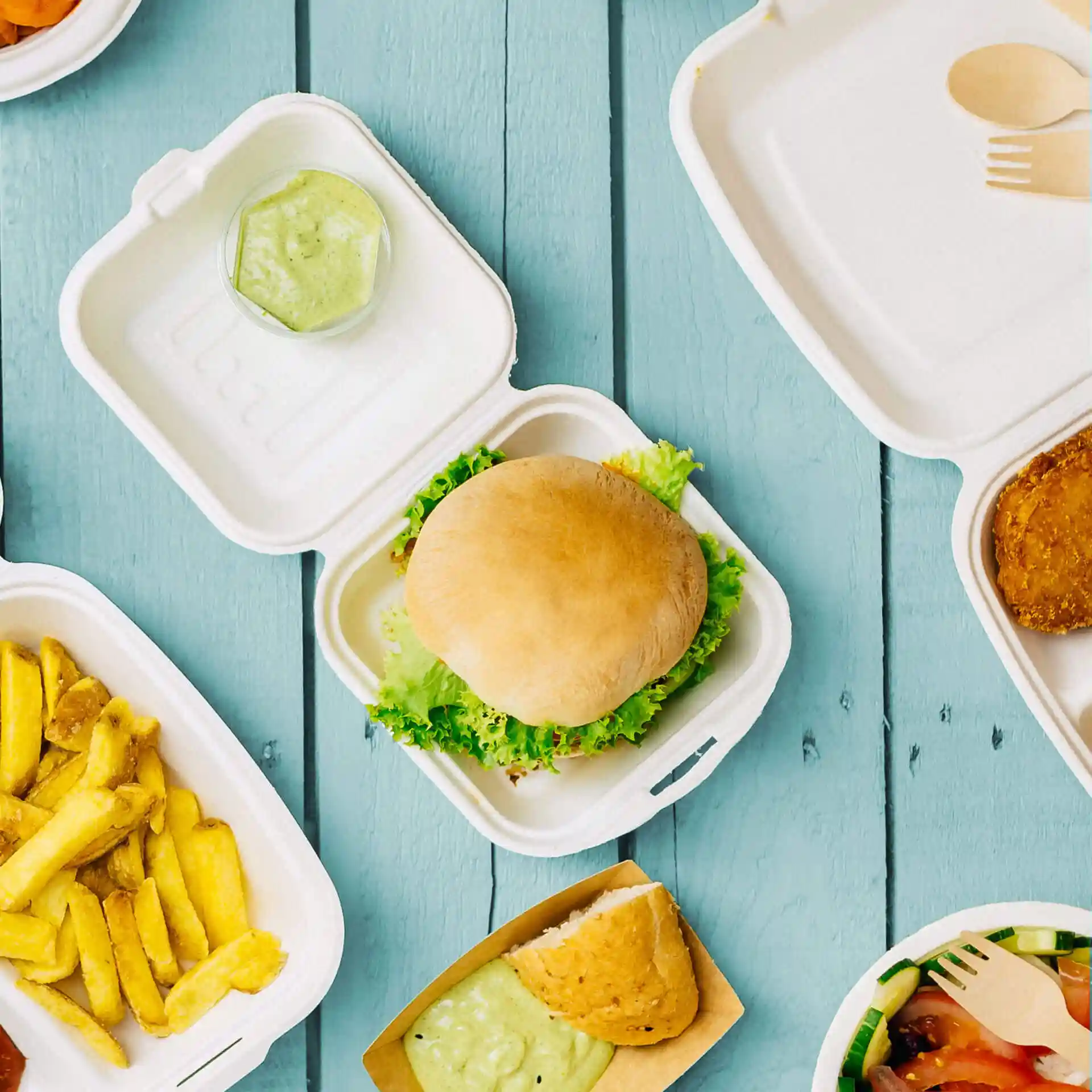 Afbeelding van een suikerrietburgerdoos met een burger met sla, een klein bakje saus en verschillende etenswaren in de buurt.