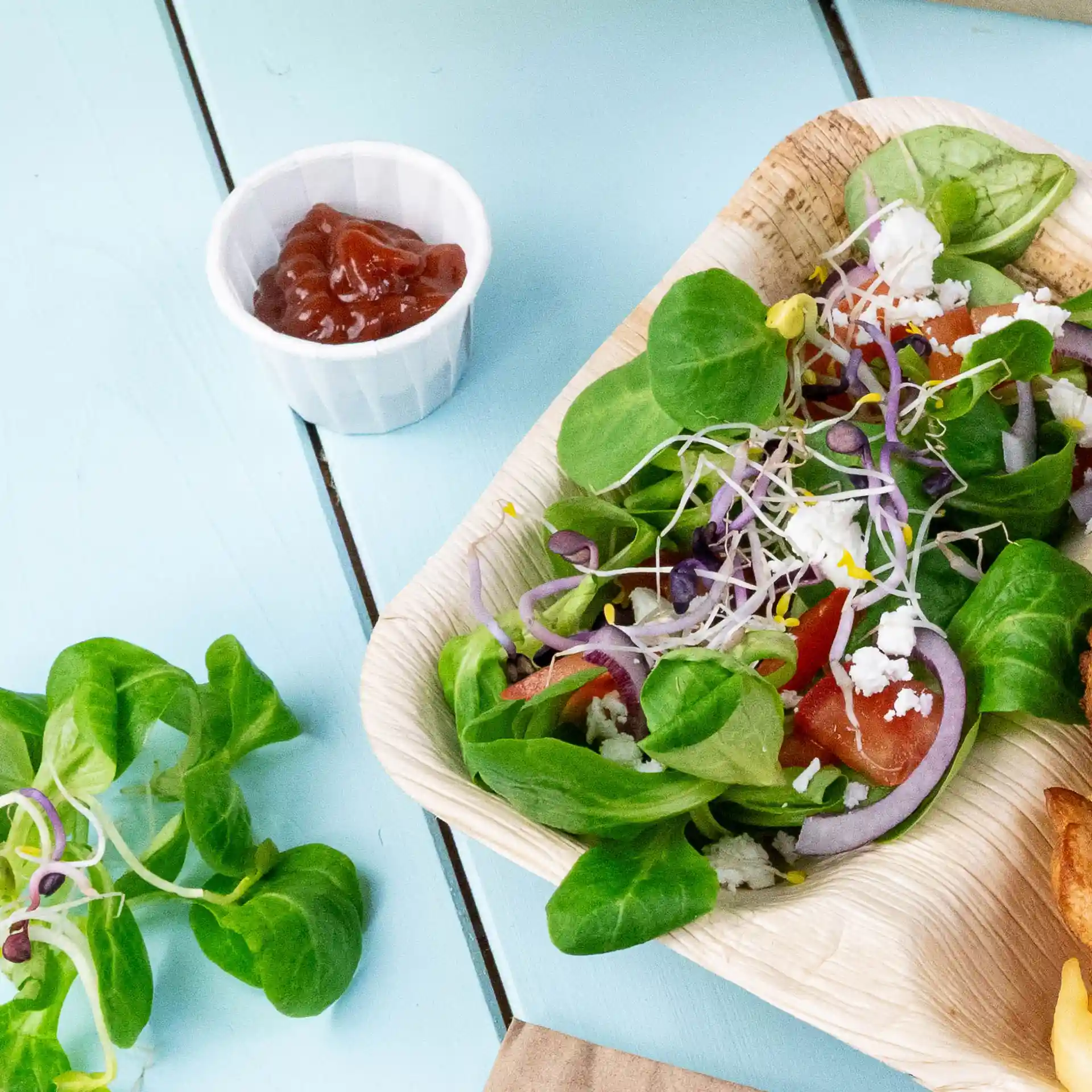 Salade met gemengde groenten, tomaten, uien en kiemen in een palmbladkom, vergezeld van een klein wit bekertje ketchup.