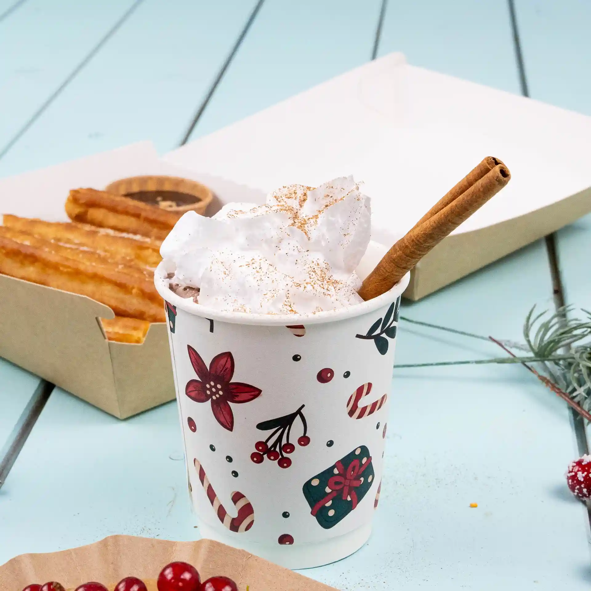 Dubbelwandige beker met winterontwerp, gevuld met slagroom en een kaneelstokje, naast churros op een blauwe ondergrond.