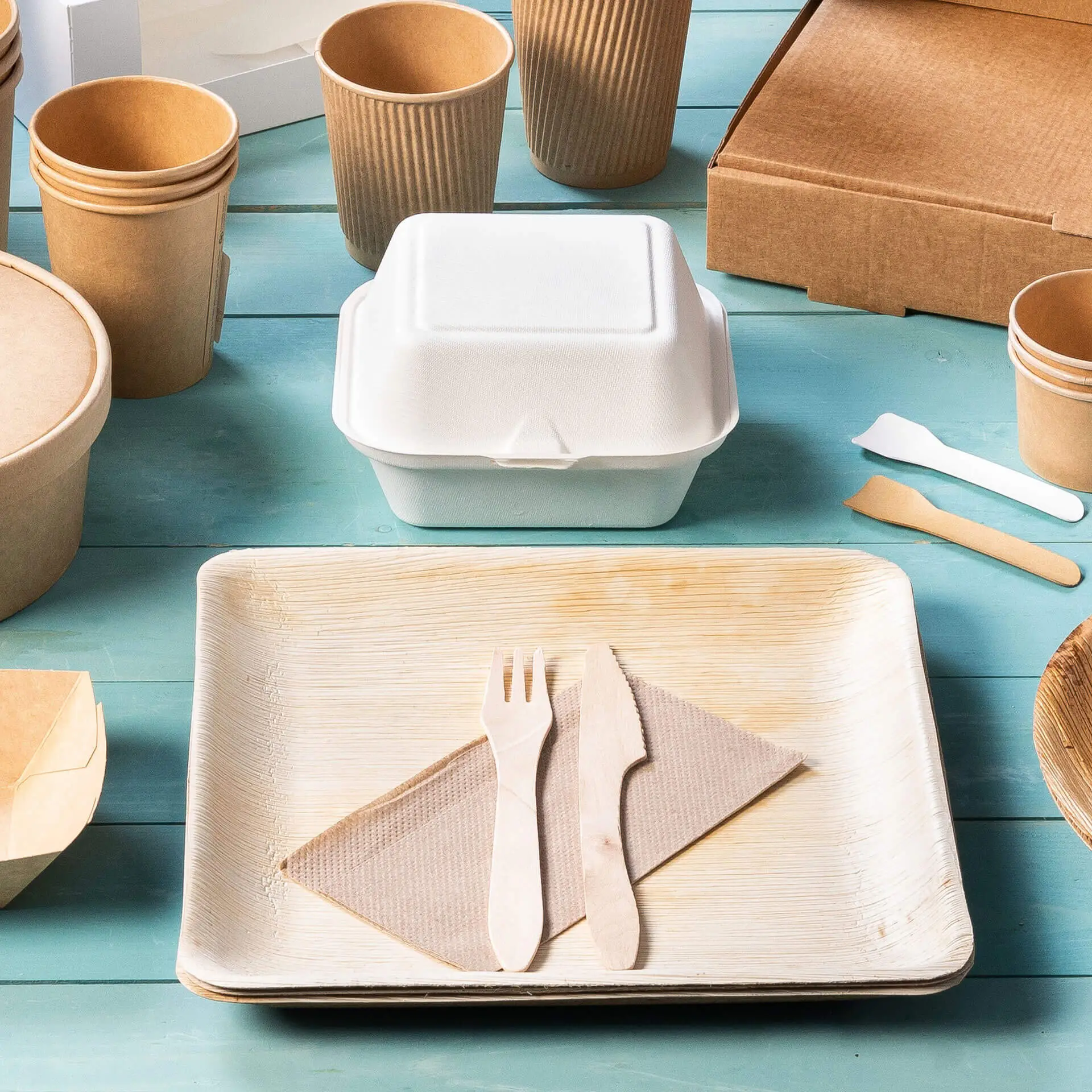 Afbeelding van een witte afhaalcontainer op een houten dienblad met een houten mes en vork op een servet, omringd door verschillende papieren bekers en doosjes.