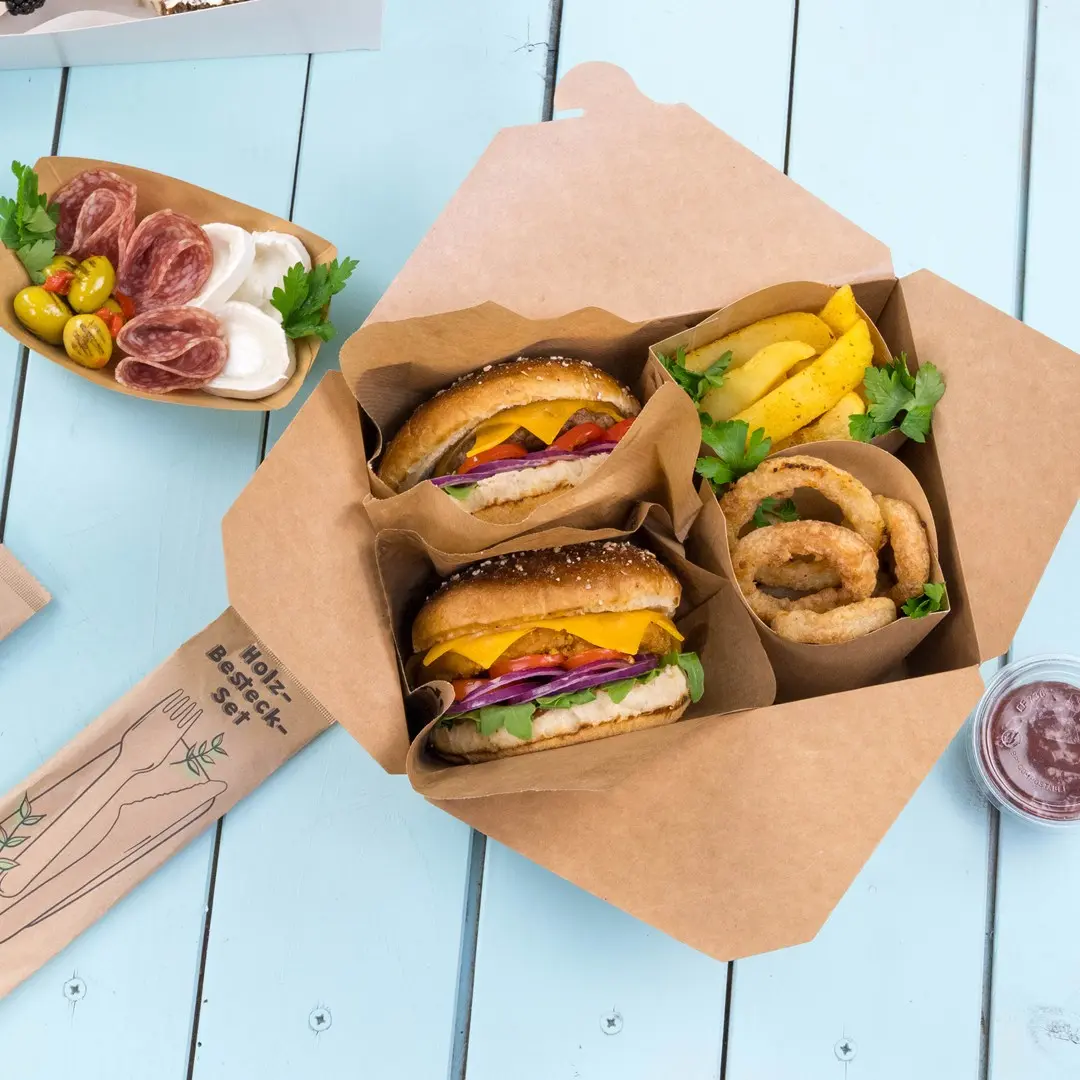 Afhaaldoos met twee hamburgers, uienringen, aardappelpartjes en een klein bakje saus op een blauwe houten ondergrond.