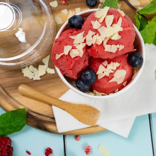 Kom met frambozensorbet, getopt met bosbessen en witte chocoladeschaafsel, houten lepel en muntblaadjes op een houten oppervlak.