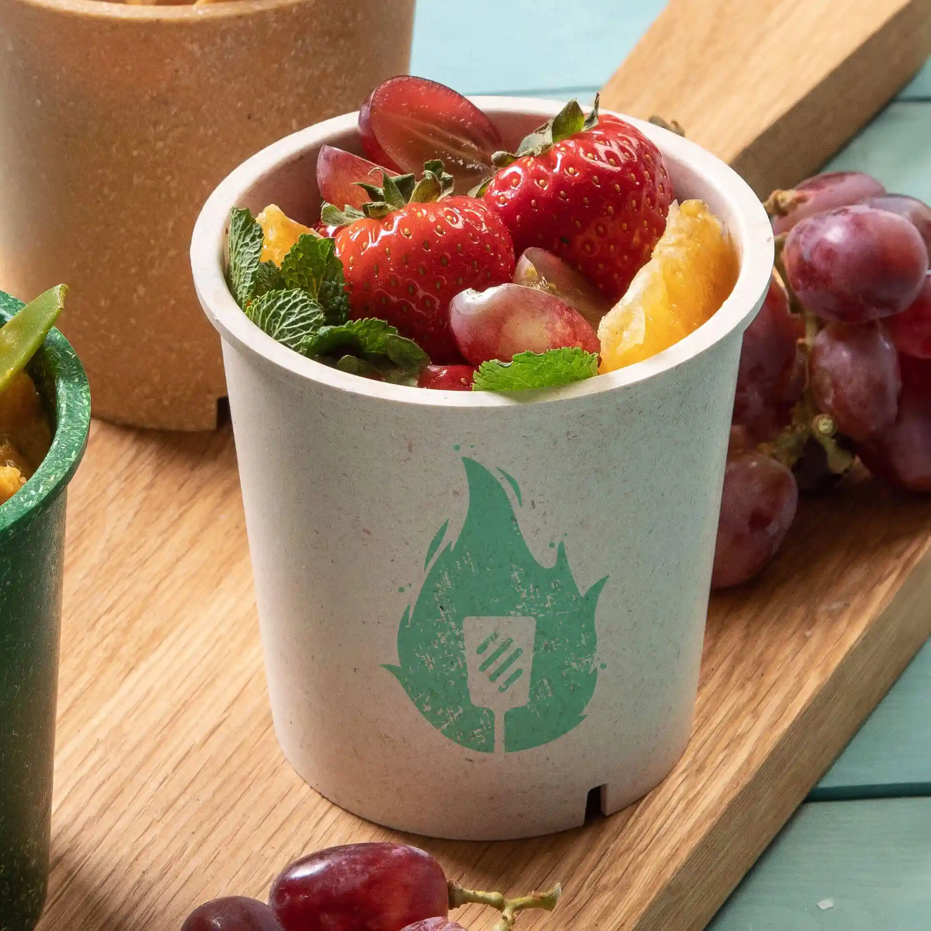 Ronde crèmekleurige beker gevuld met aardbeien, druiven en muntbladeren, met een groen vlam symbool.