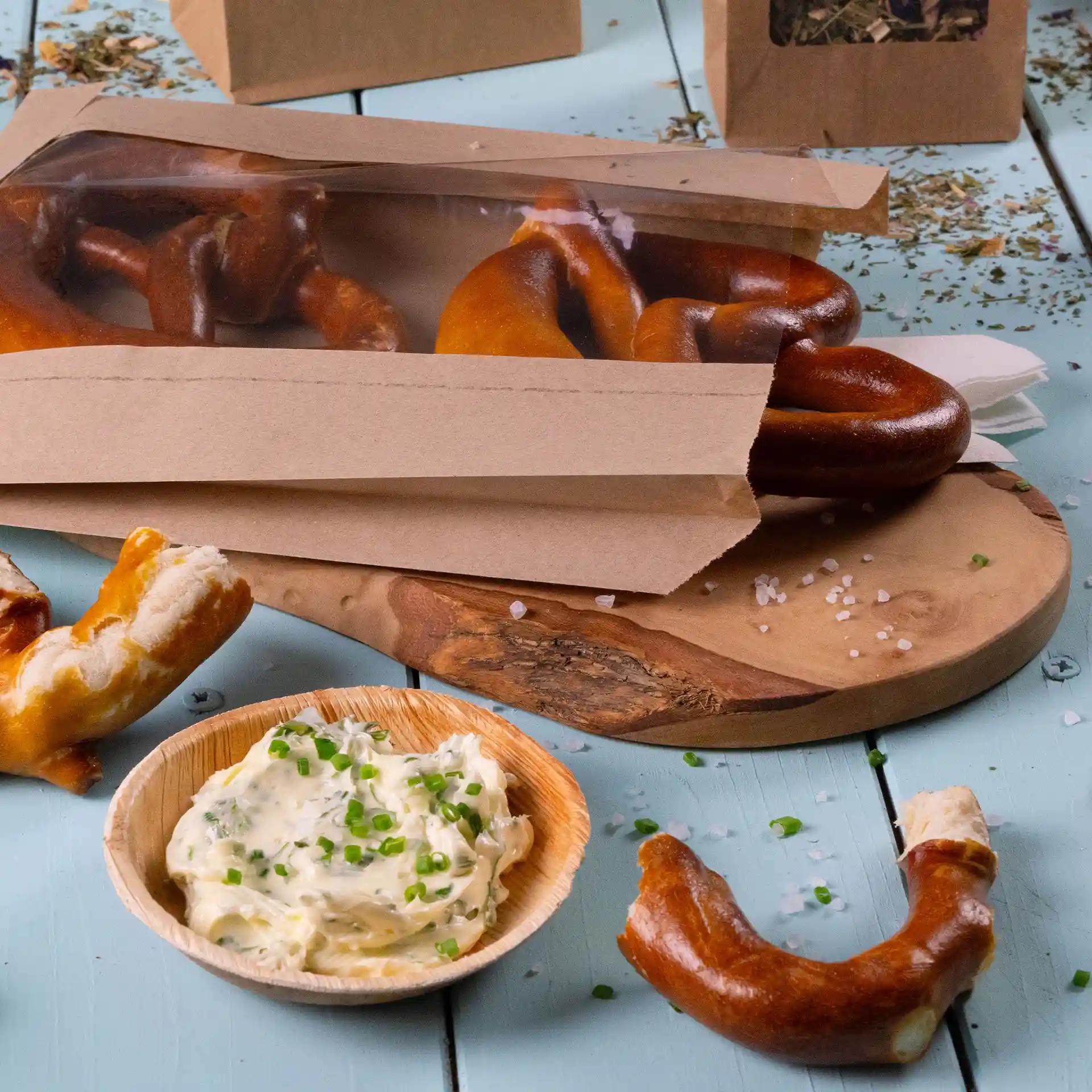 Bruine platte papieren zak met een venster met pretzels, naast een houten bord met romige dip en bieslook.