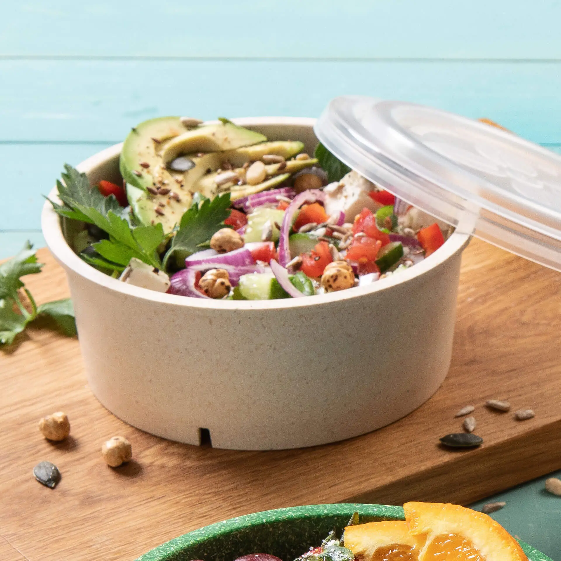 Een saladeschaal met gemengde groenen, avocado-helften, tomaten en zaden; doorzichtig deksel erop, houten achtergrond.