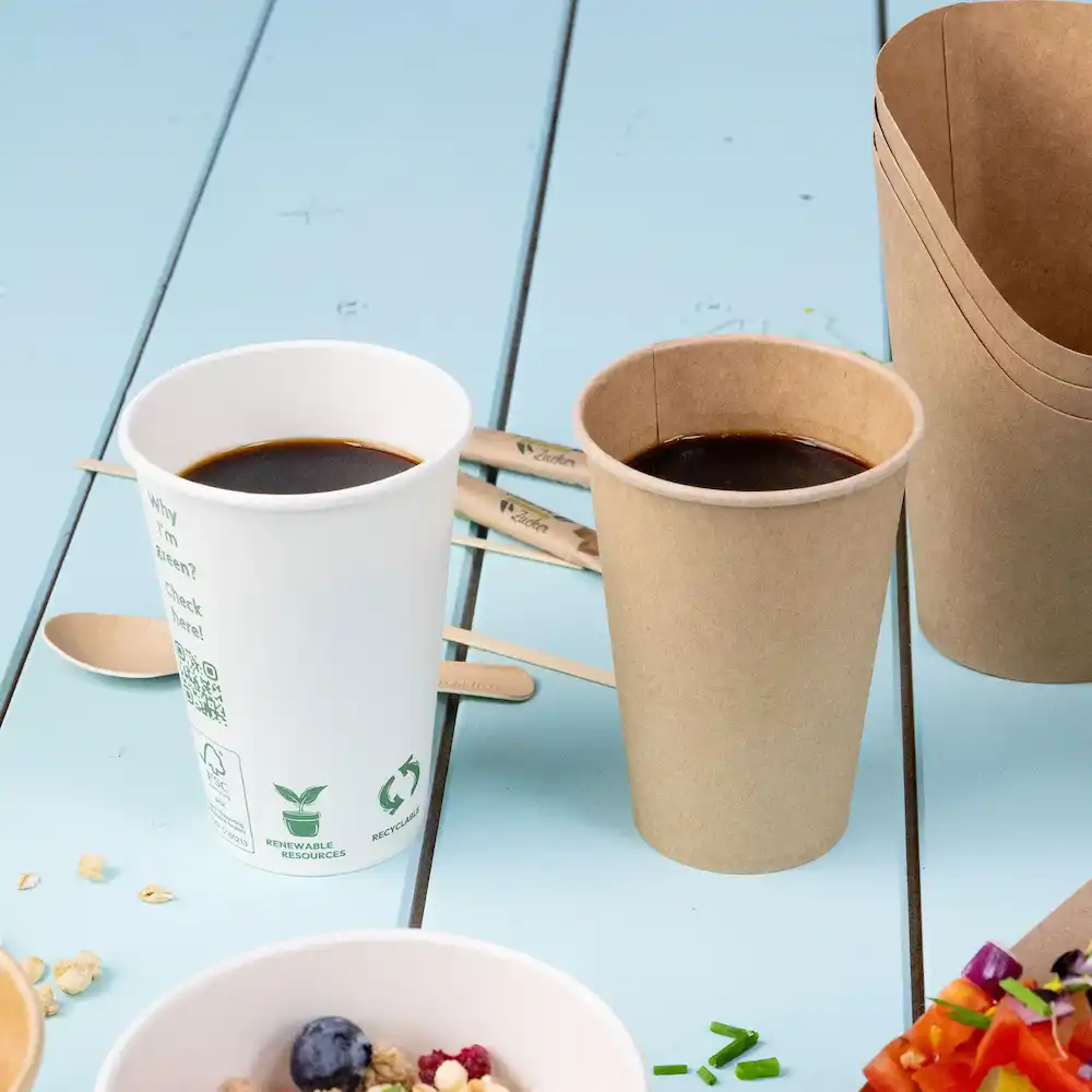 Twee cups met koffie, de ene wit en de andere bruin, naast houten roerstaafjes op een blauwe ondergrond.