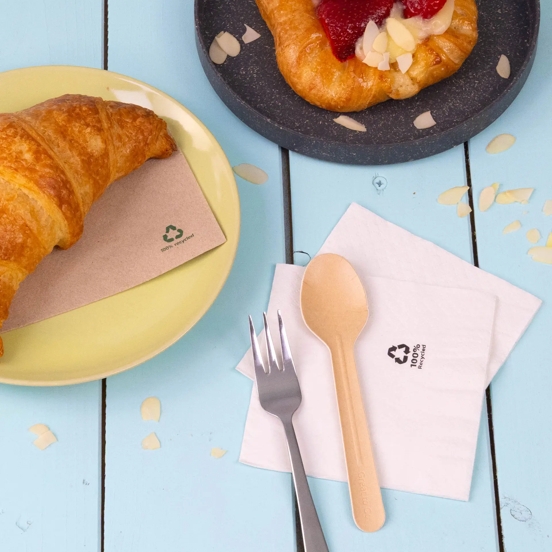 Een croissant op een gele bord naast een witte 2-laags gerecycleerde papieren servet en een milieuvriendelijke houten lepel en vork.