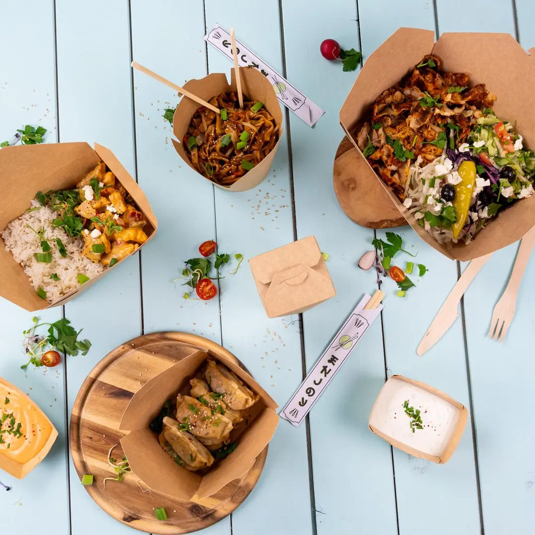 Diverse take-away voedselcontainers met noedels, rijst, salade en dumplings op een blauwe houten ondergrond.