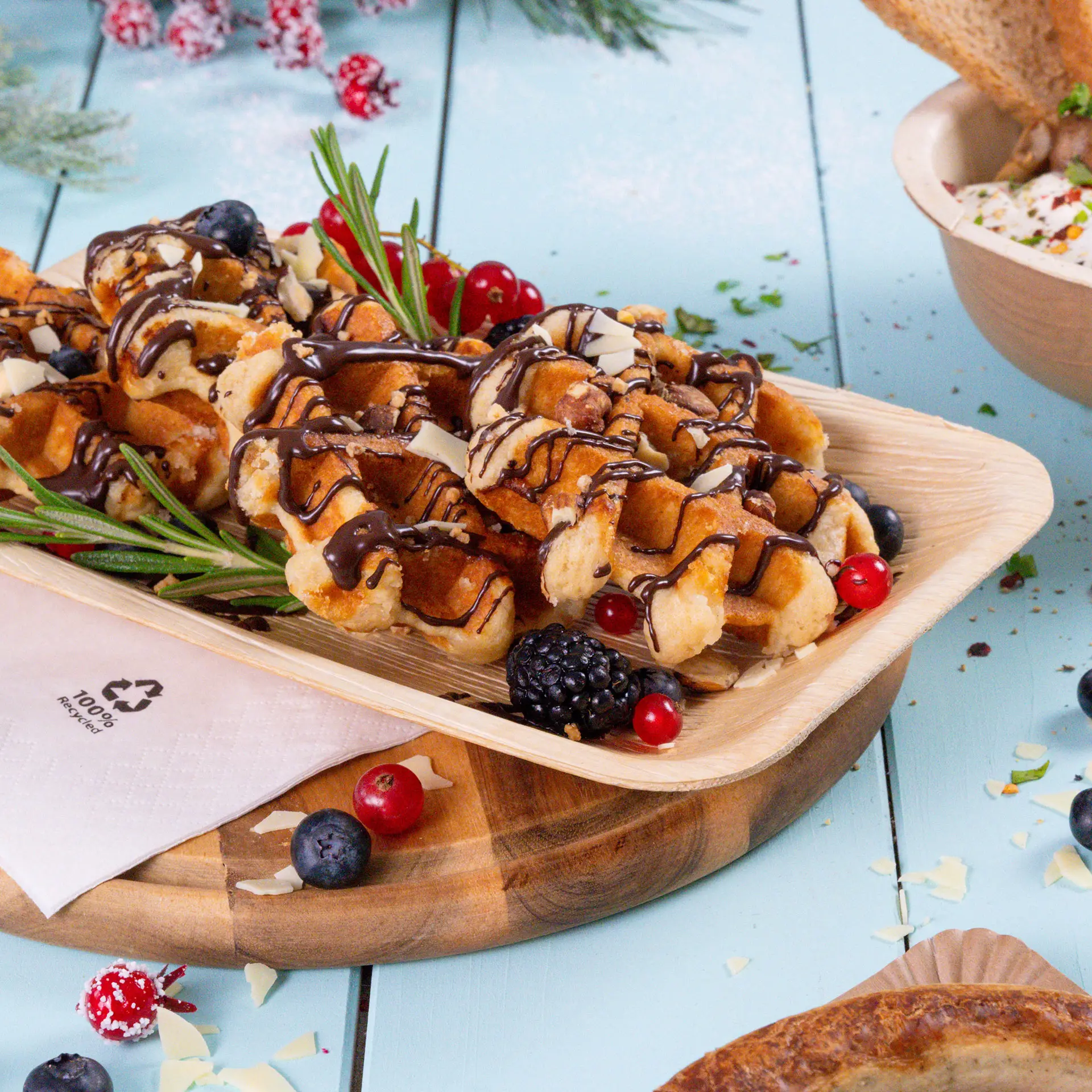 Bord met wafels, besprenkeld met chocolade, gegarneerd met bessen en rozemarijn, op een wegwerp bord van palmblad.