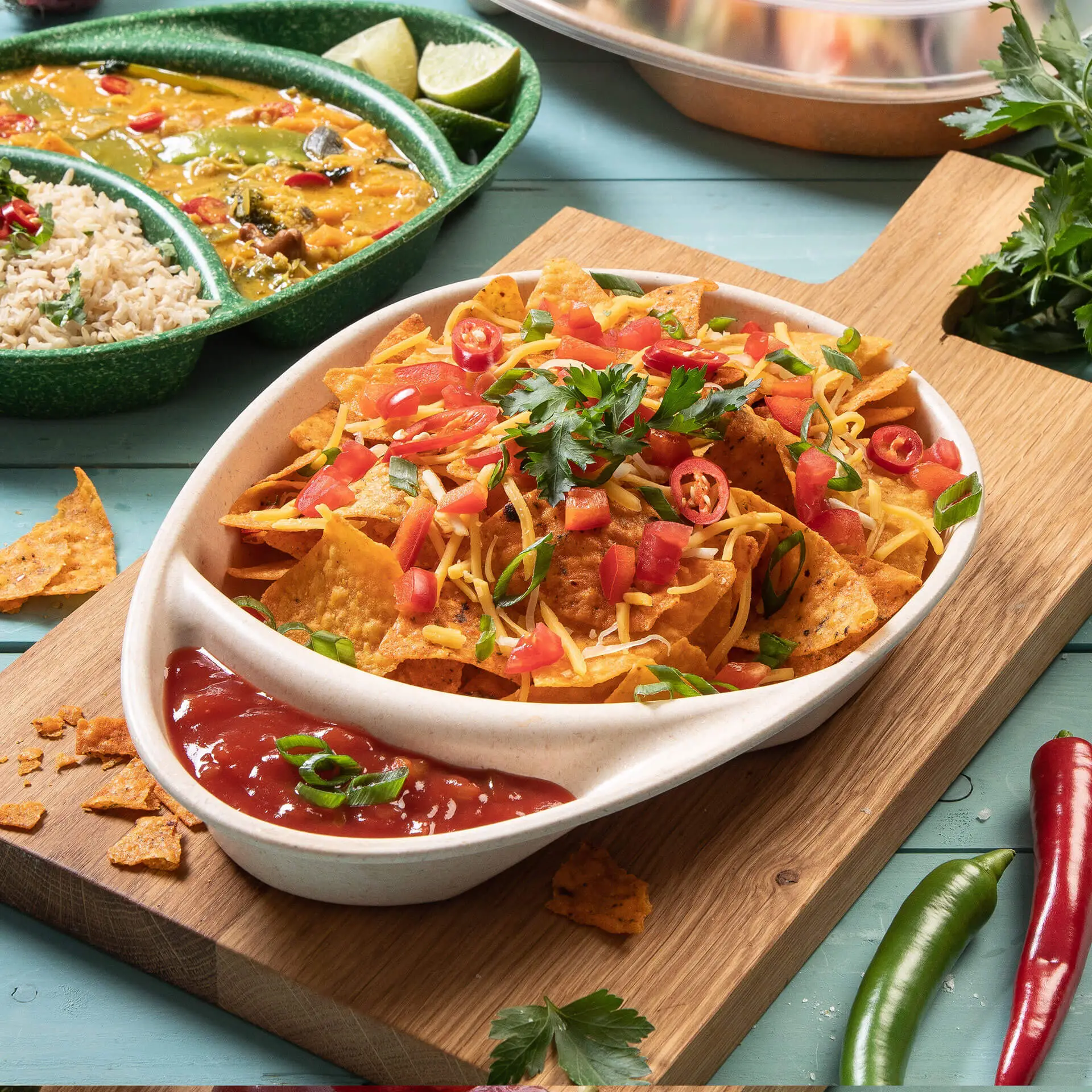 Een kom nacho's belegd met kaas, tomaten en koriander; geserveerd met salsa op een houten schaal. Extra gerecht op de achtergrond.