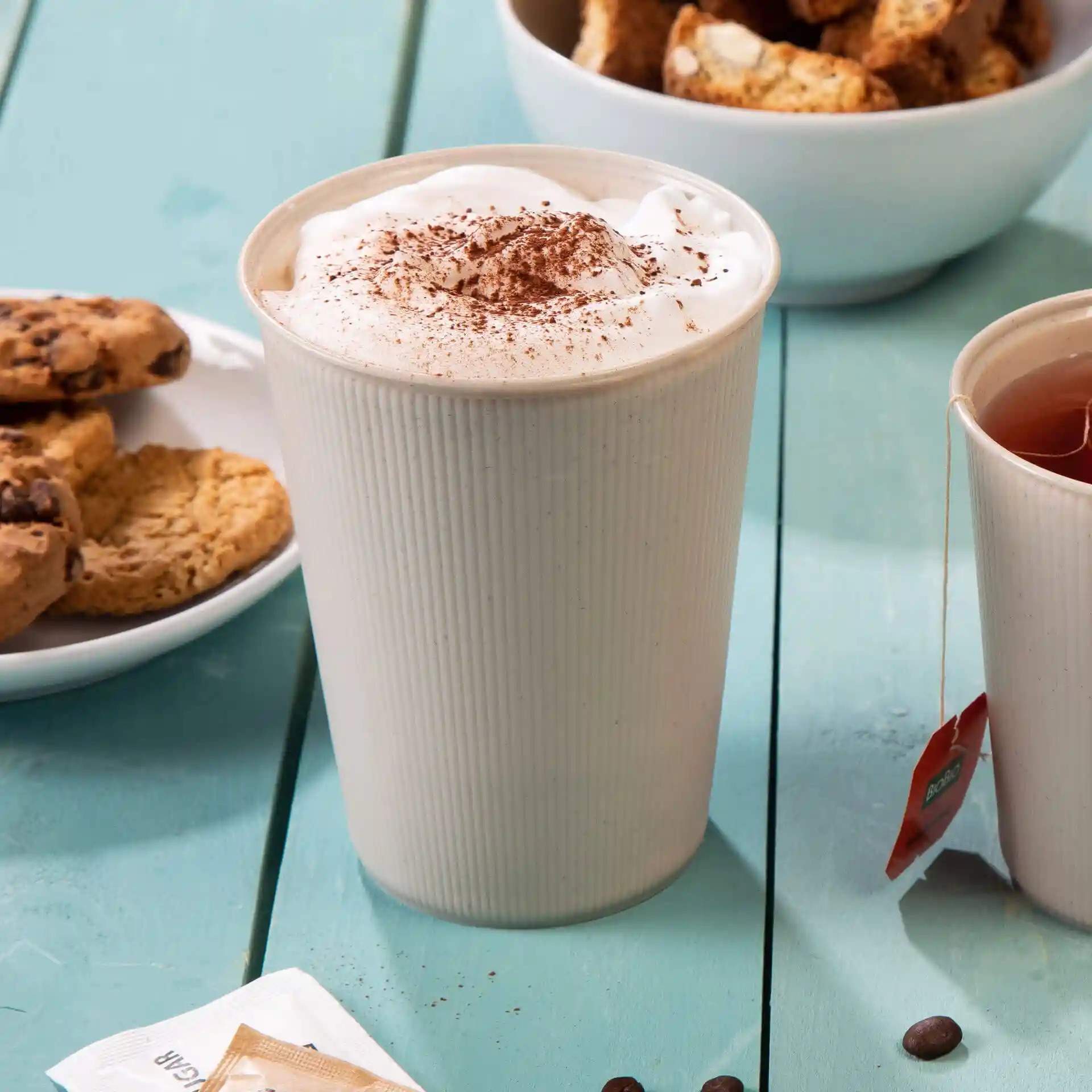Crème-witte ribbelbeker, herbruikbaar, gevuld met slagroom en cacao, met koekjes en thee op een blauwe tafel.