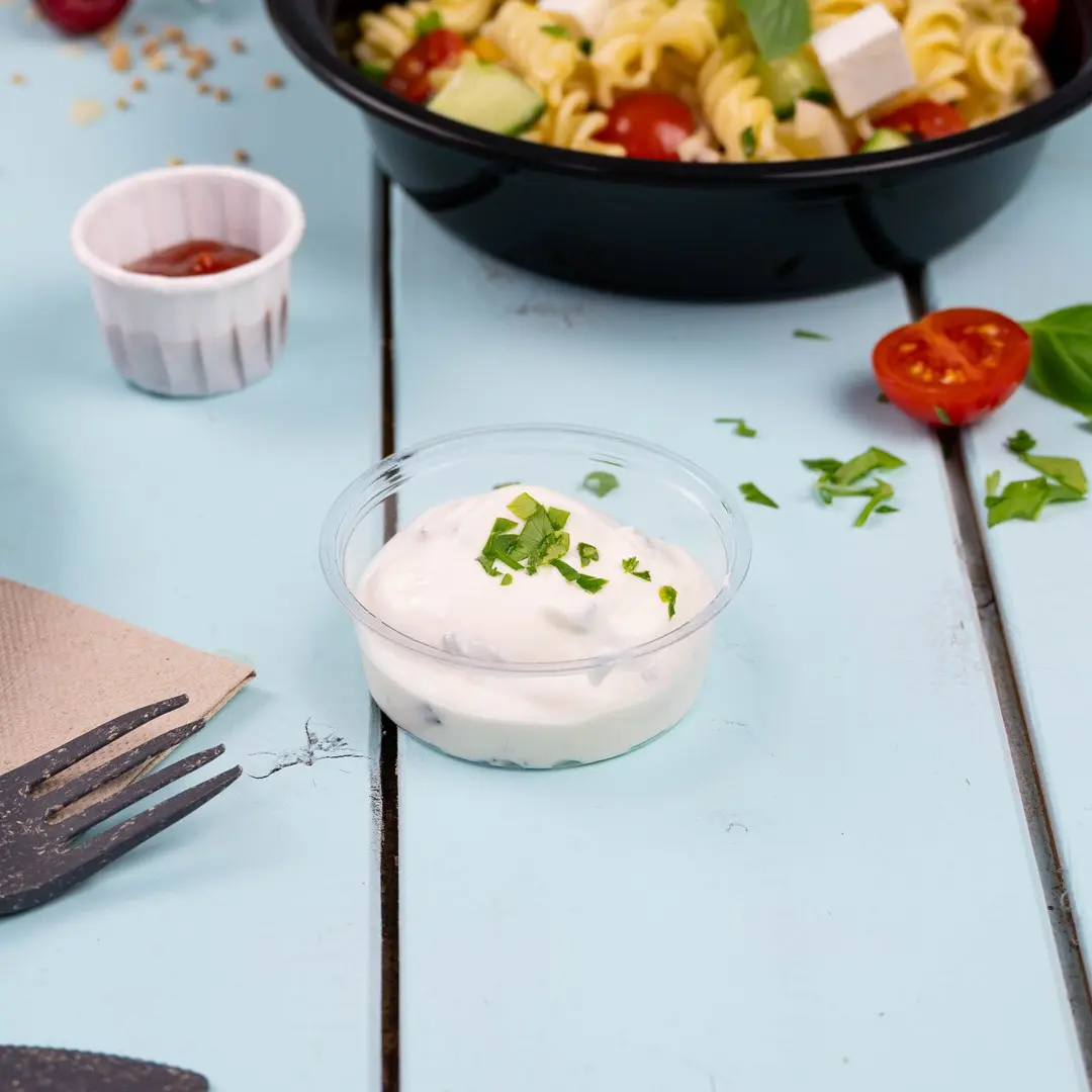 Heldere plastic beker met romige saus, getopt met gehakte kruiden, op een lichtblauwe houten ondergrond.
