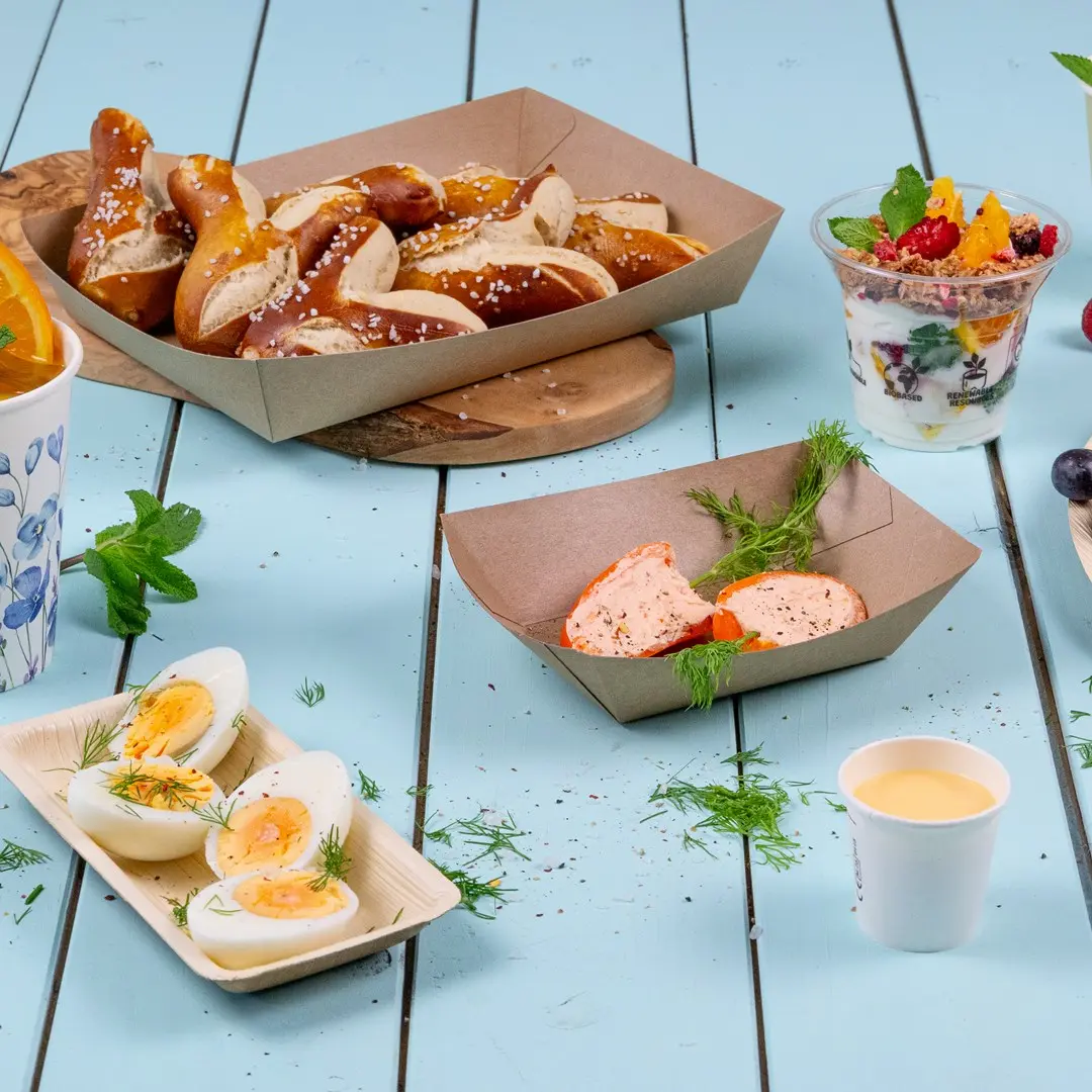 Diverse snacks, waaronder pretzels, gekookte eieren, yoghurtparfait en een klein bekertje saus op een blauwe houten ondergrond.