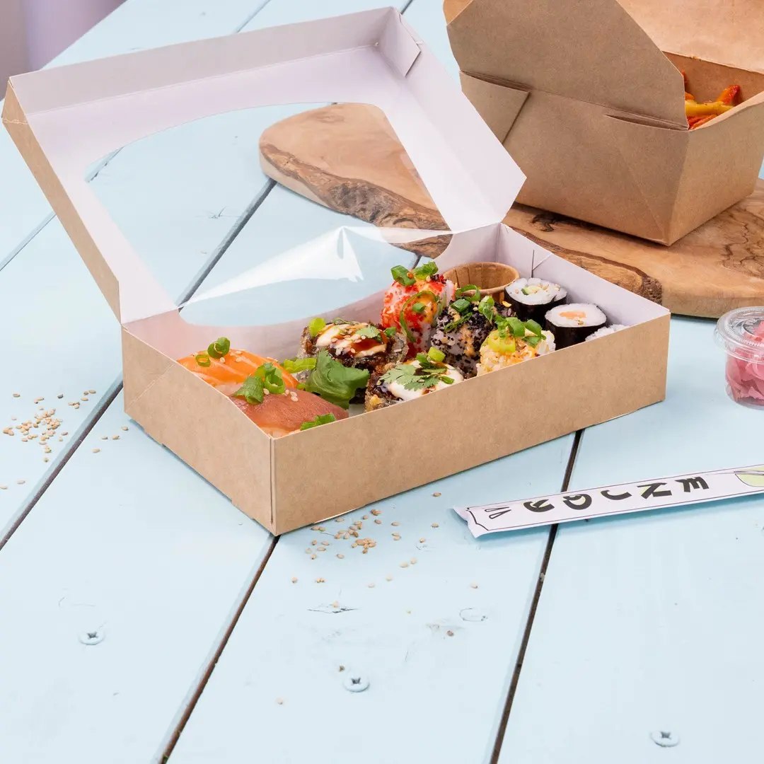 Sushi-doos met verschillende sushirollen, zalm en garnituren, geplaatst op een lichtblauwe houten ondergrond.
