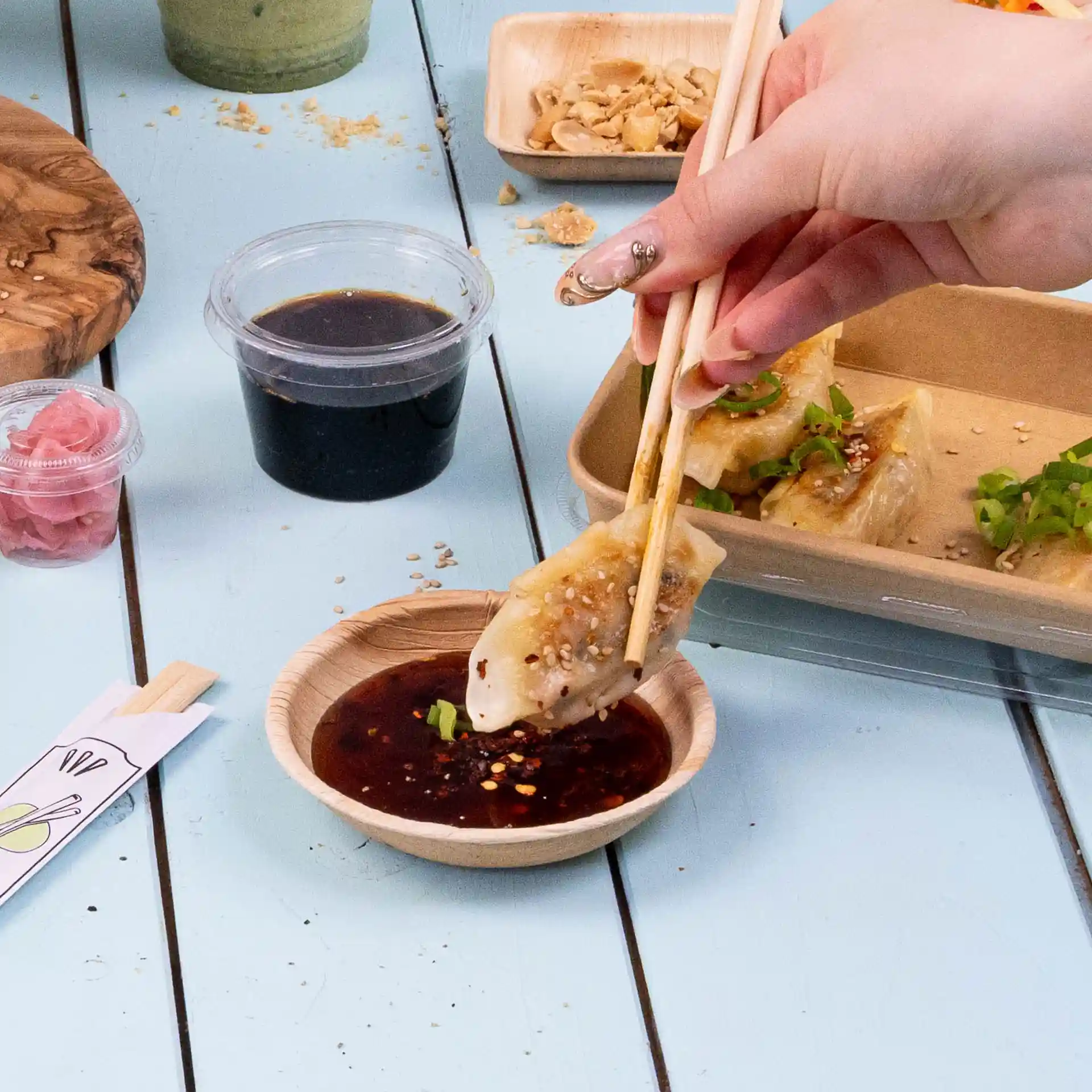 Een hand met chopsticks doopt een dumpling in een ronde palmbladkom met donkere saus.