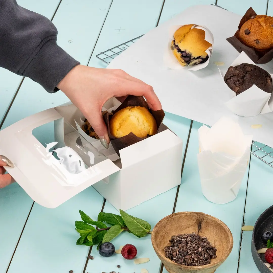 Hand plaatst een muffin in een witte cupcake-doos met verschillende gebakken goederen en ingrediënten op een blauwe ondergrond.