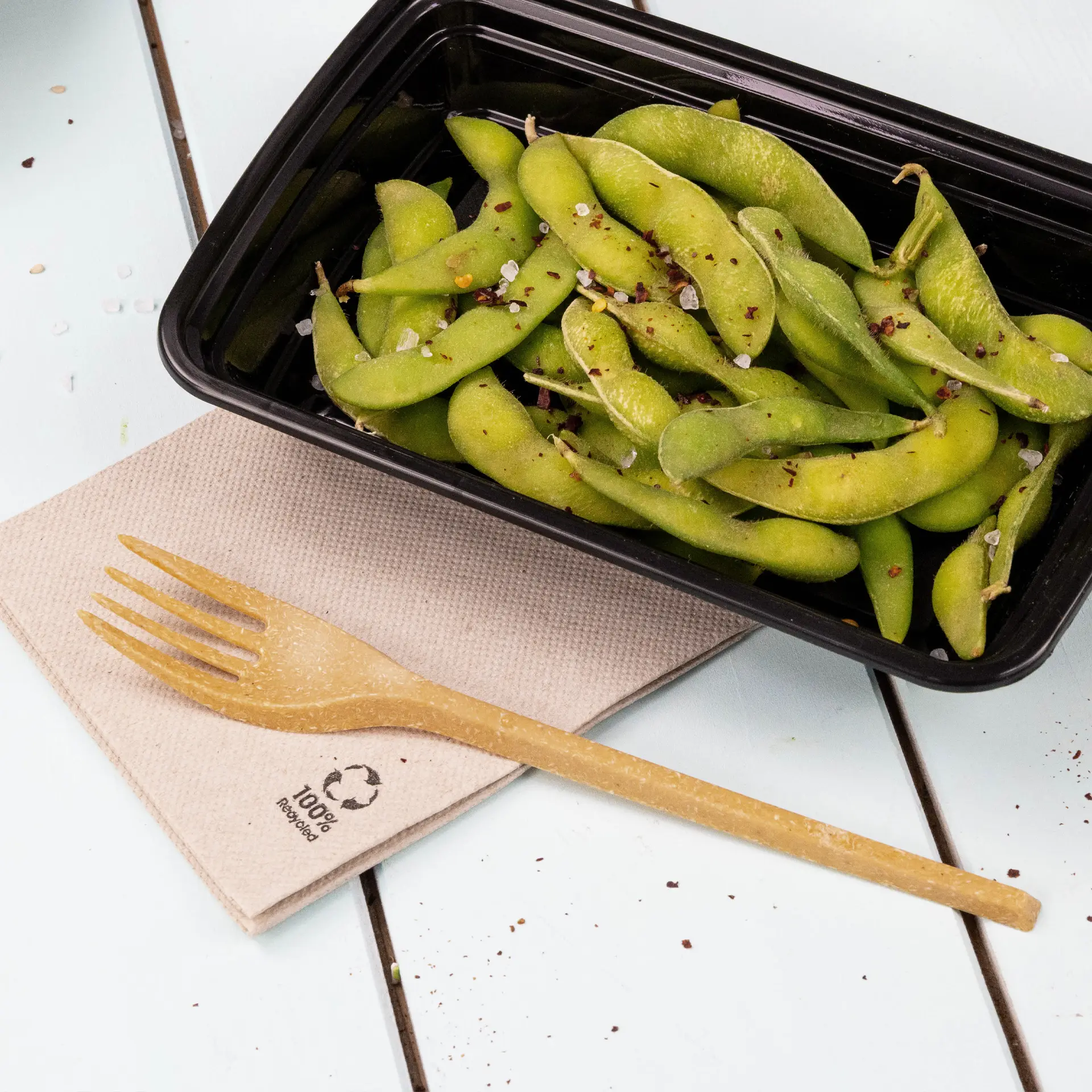 Een zwarte container gevuld met groene edamame, gegarneerd met specerijen, vergezeld van een biologisch afbreekbare vork op een gerecycled papieren servet.
