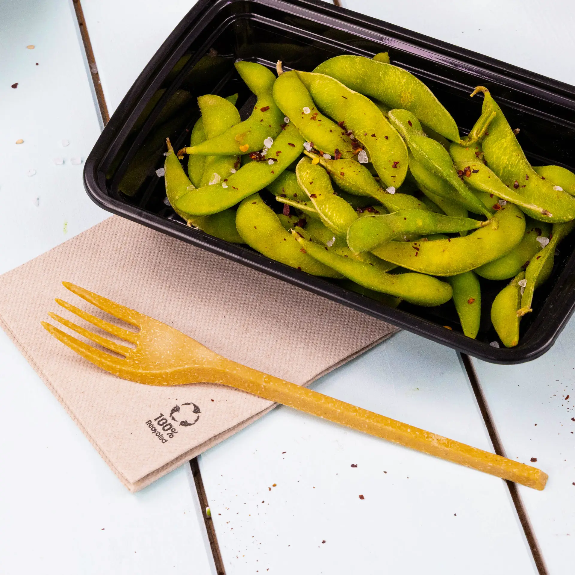 Een zwarte container gevuld met groene edamame, gegarneerd met specerijen, vergezeld van een biologisch afbreekbare vork op een gerecycled papieren servet.