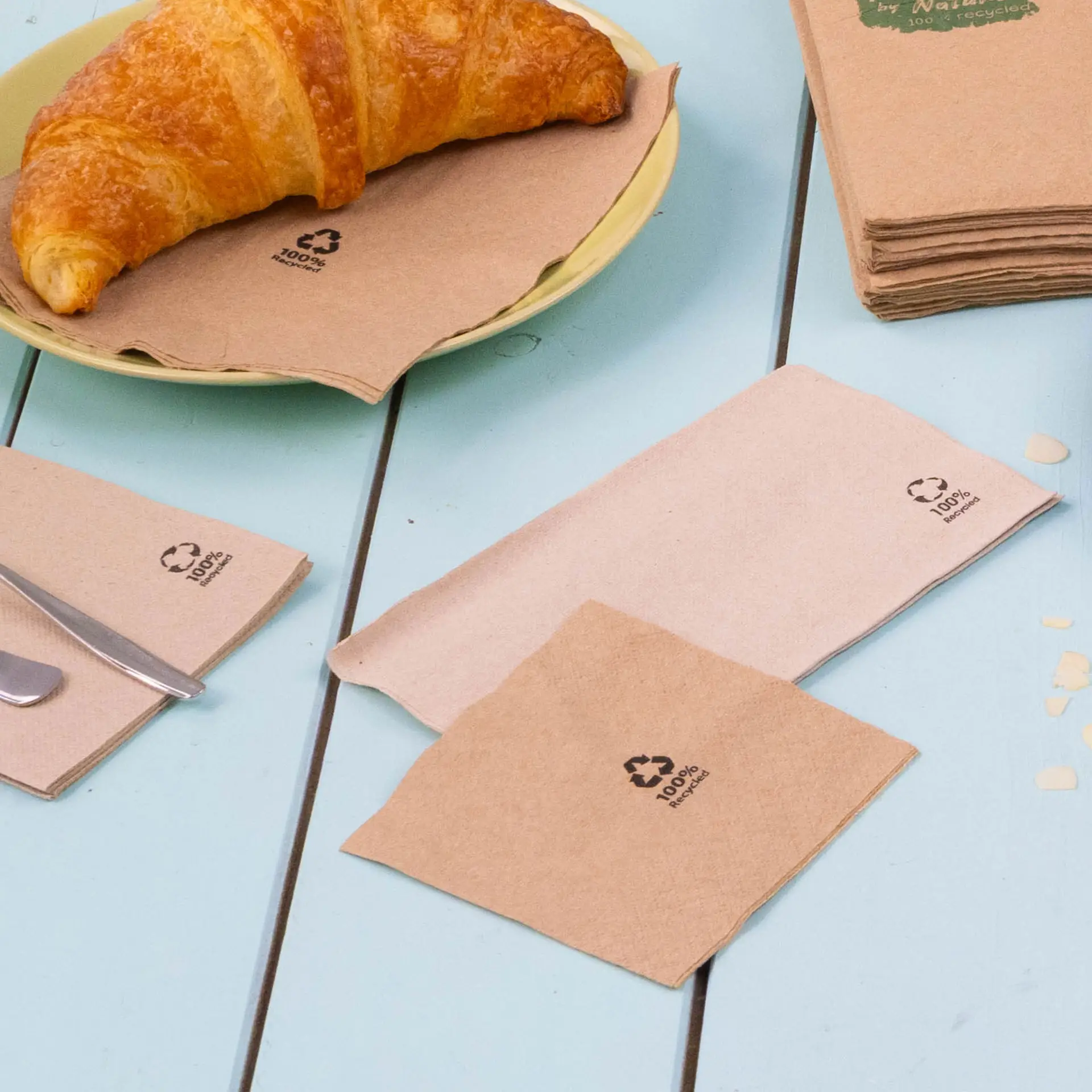 Afbeelding van bruine, biologisch afbreekbare cocktailservetten op een blauwe tafel, met een croissant op een bord, met recycling-symbolen.