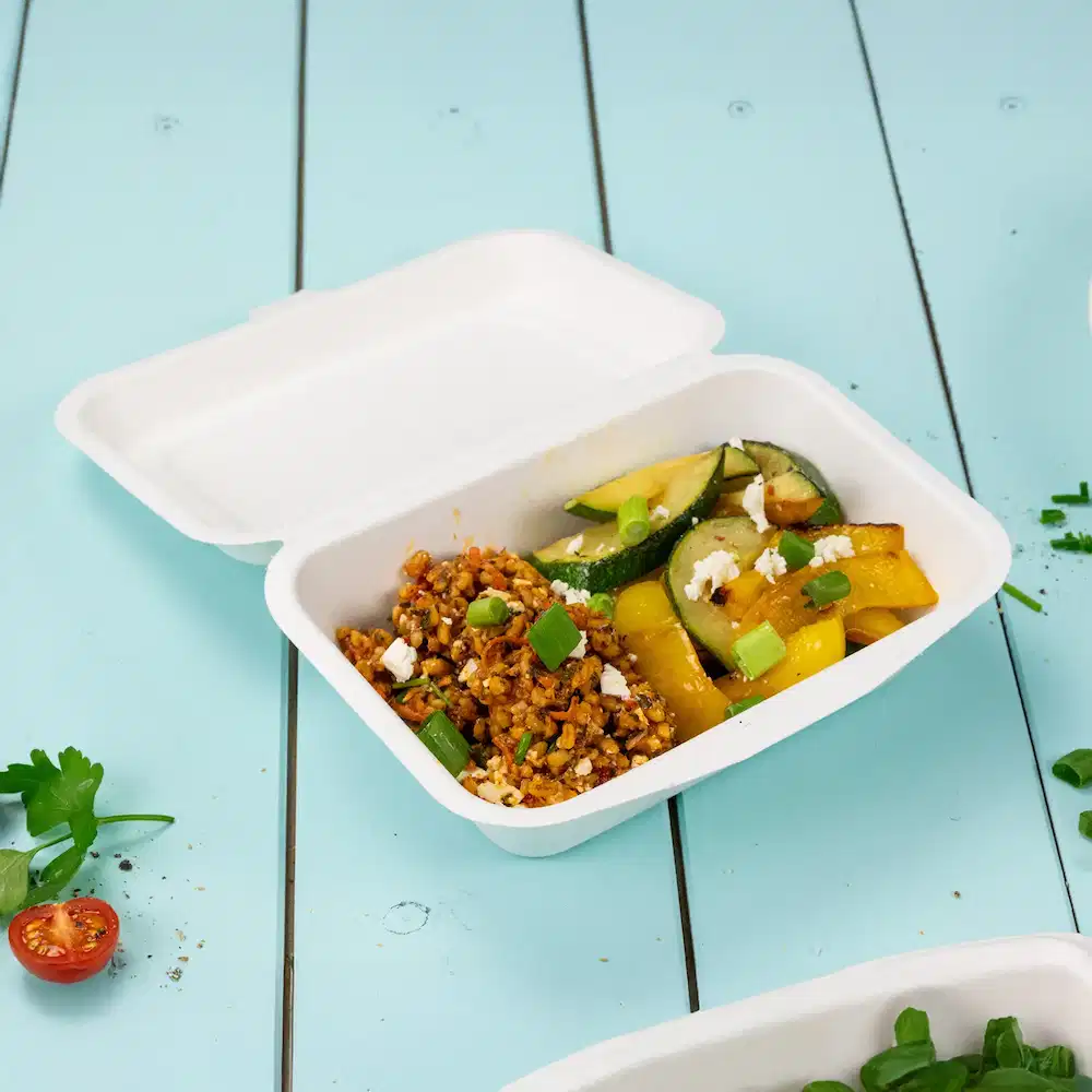 Witte voedselcontainer met een mix van gekookte granen, courgette, paprika en gegarneerd met lente-ui en verkruimelde kaas.