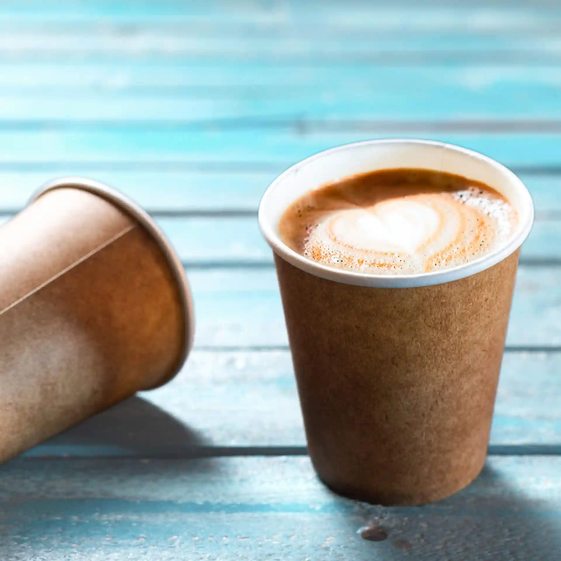 Bruine papieren beker met een witte binnenkant, gevuld met koffie en hartvormige latte-art, op een blauwe houten ondergrond.