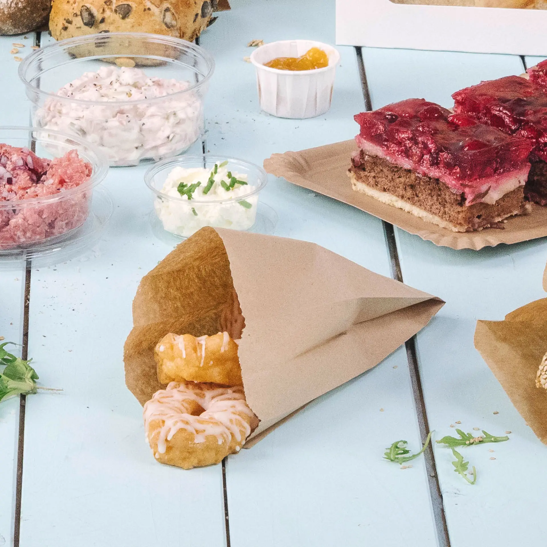 Bruine papieren zak met twee gedeeltelijk zichtbare, geglazuurde donuts; verschillende voedselcontainers op de achtergrond.