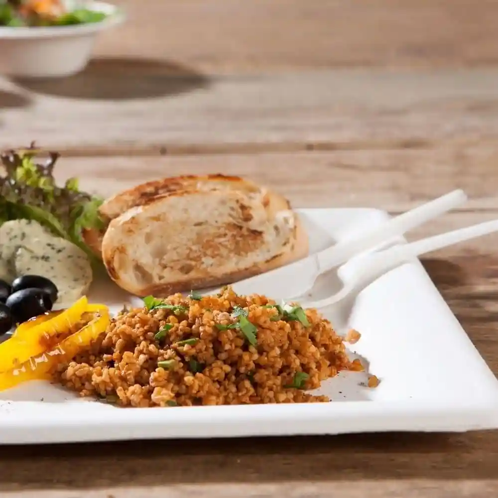 Bord met bulgur, gegrilde groenten, olijven, een stuk brood en een salade op een houten tafel.