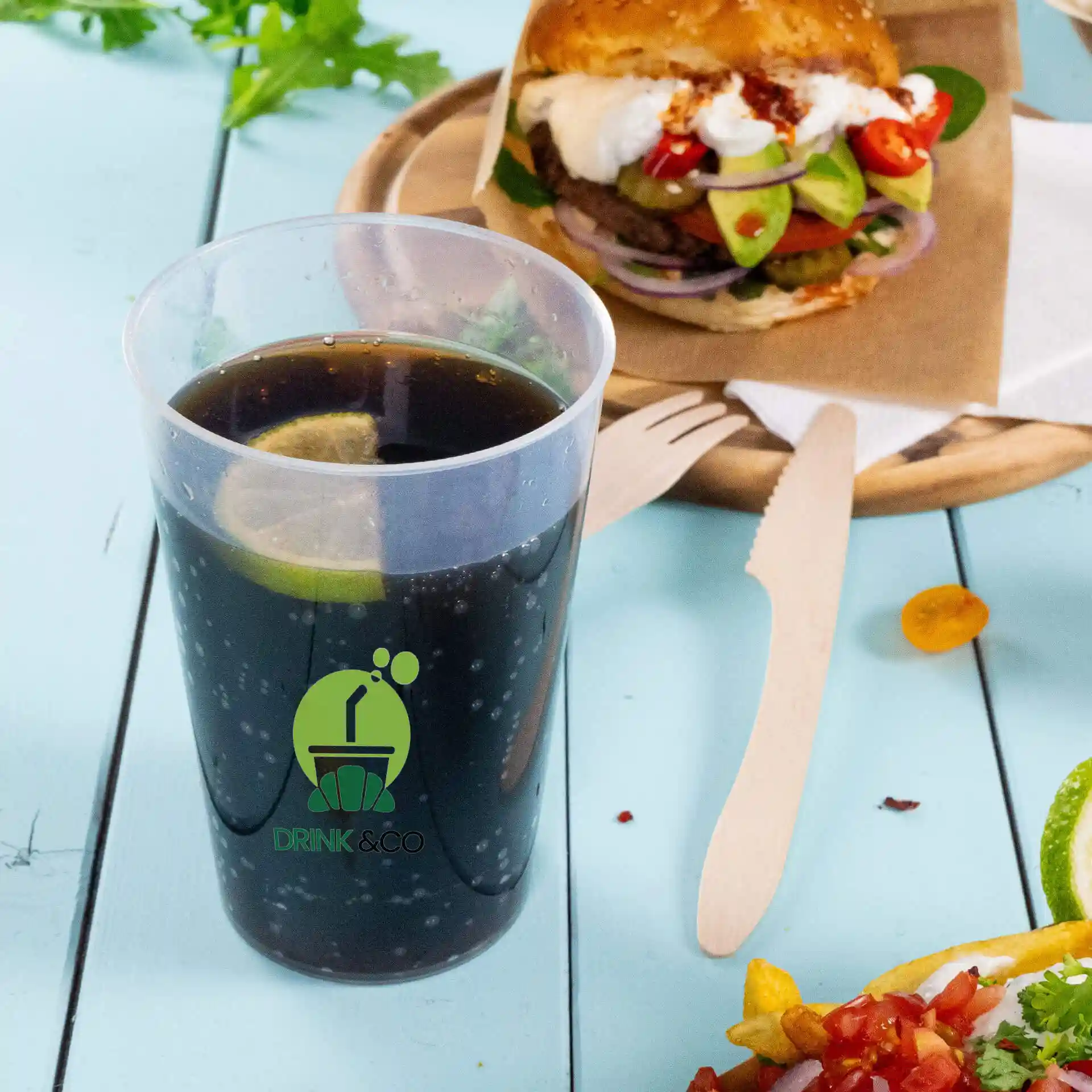 Duidelijke herbruikbare beker met logo, 500 ml capaciteit, gevuld met een donkere drank en een limoenpartje, naast een burger en salade geplaatst.