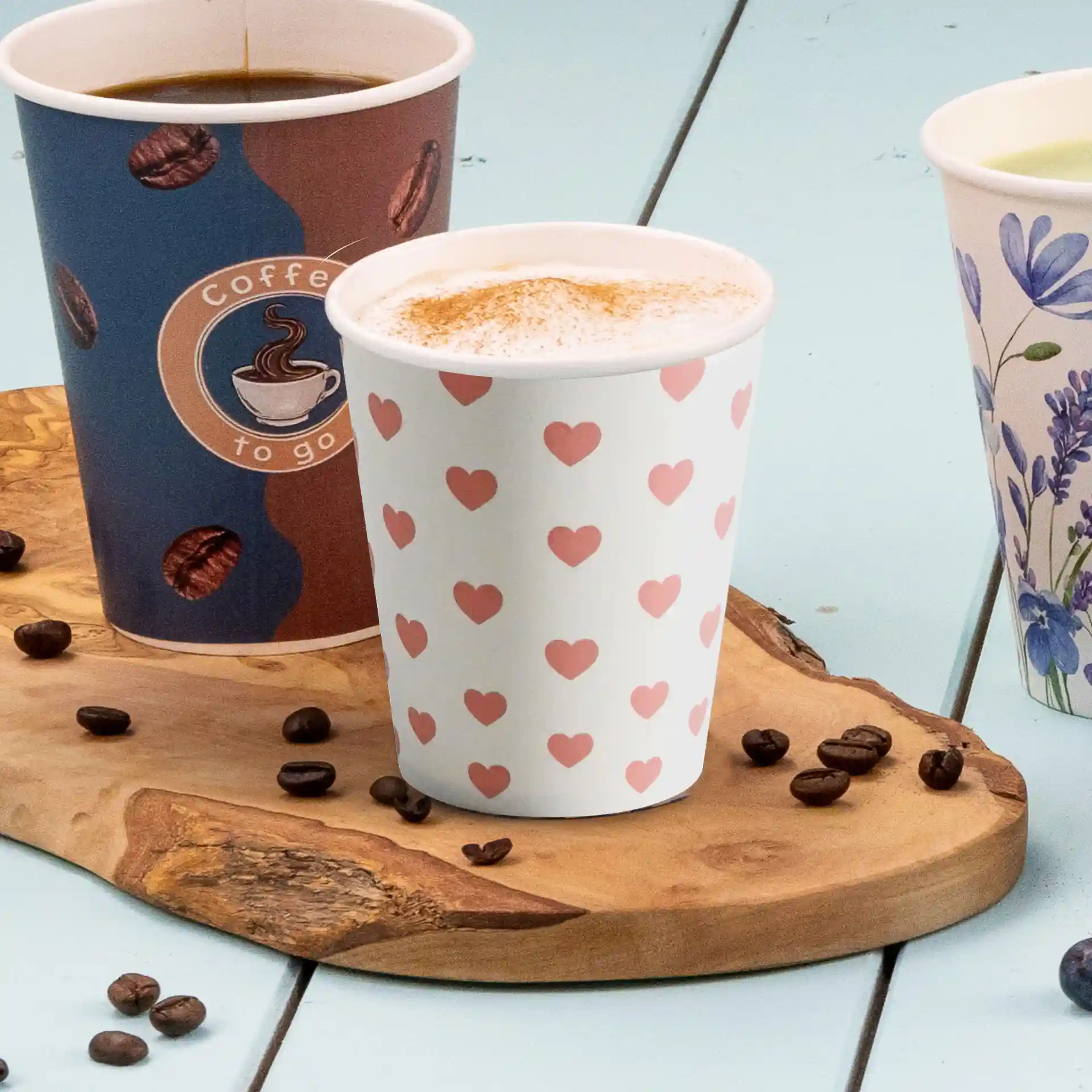 Drie papieren bekers op een houten plank, één met een hartpatroon, één met een koffielogo en één met een bloemenontwerp.