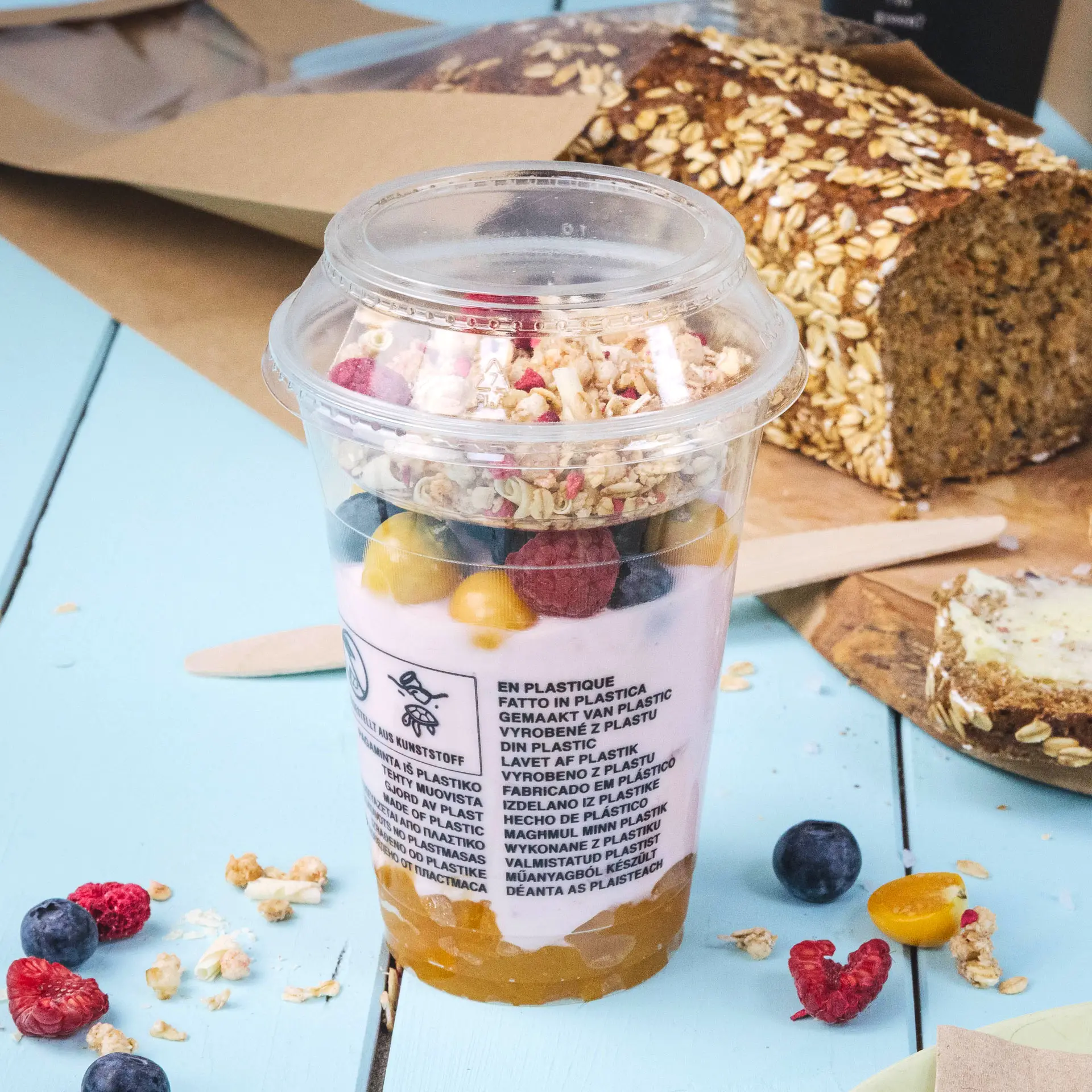 Een doorzichtig PLA smoothiebeker gevuld met yoghurt, granola en fruit, geplaatst op een blauwe ondergrond naast een brood.