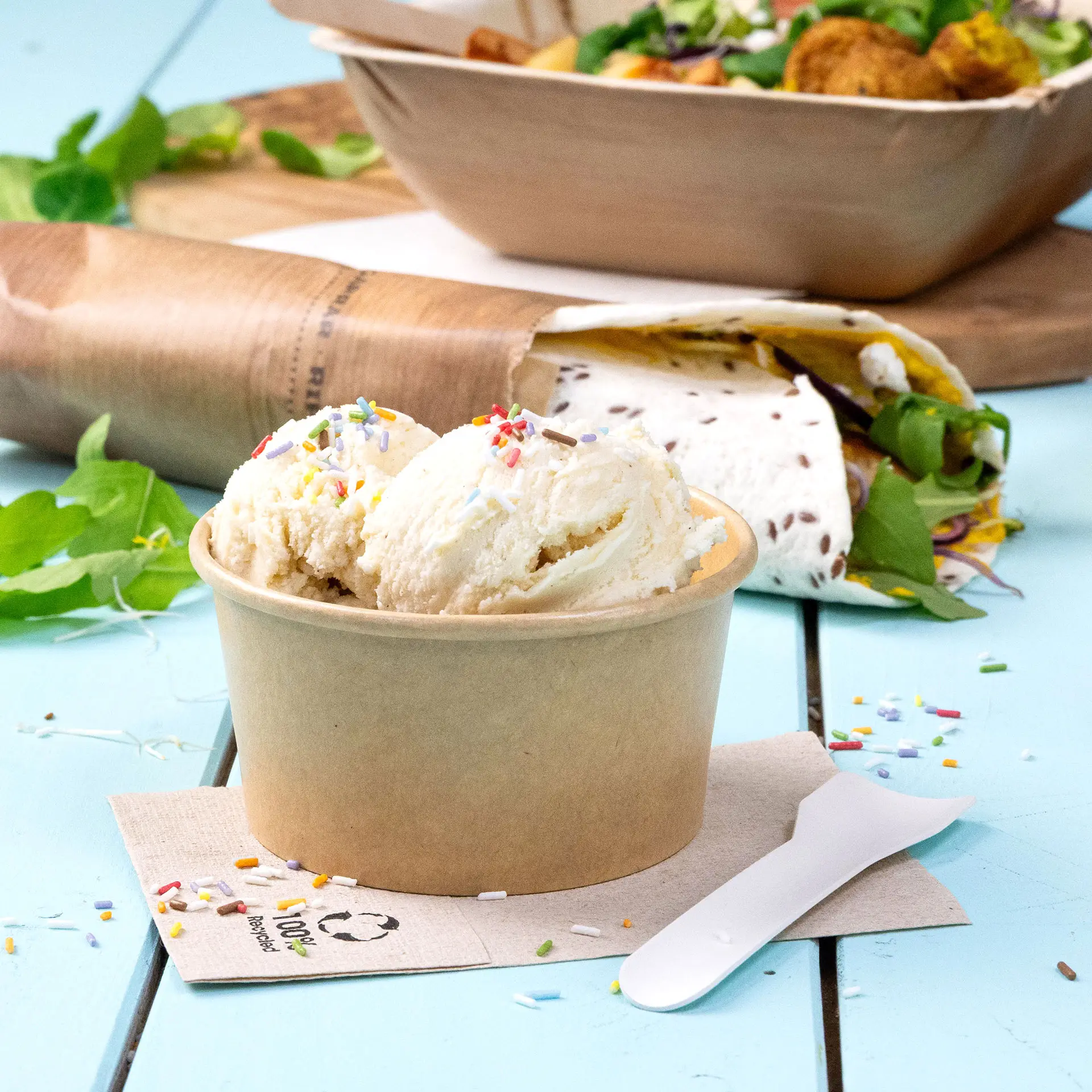 Bruine papieren ijscoupe met twee bollen ijs, gegarneerd met kleurrijke sprinkles; een lepel en een servet ernaast.