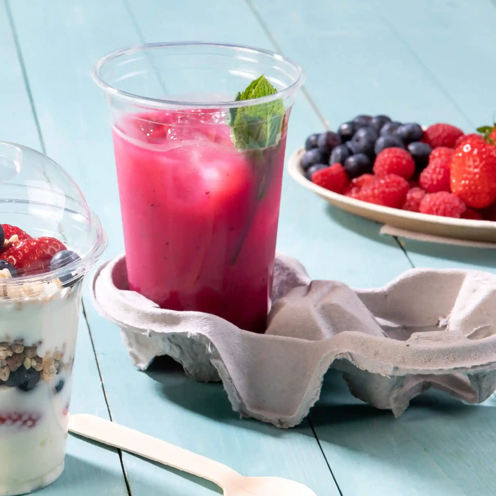 Heldere 16 oz rPET-beker gevuld met een roze drankje en munt, geplaatst in een grijze eierdoos met een yoghurtparfait erbij.