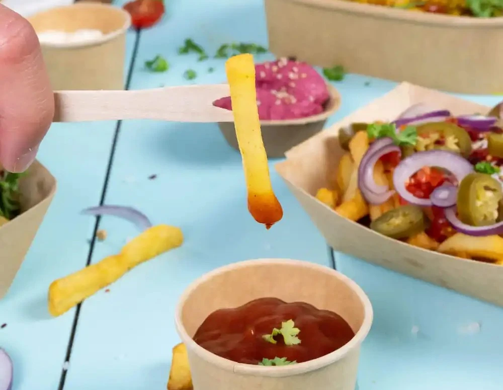 Een hand houdt een houten stok met een friet boven een beker ketchup op een blauwe ondergrond.