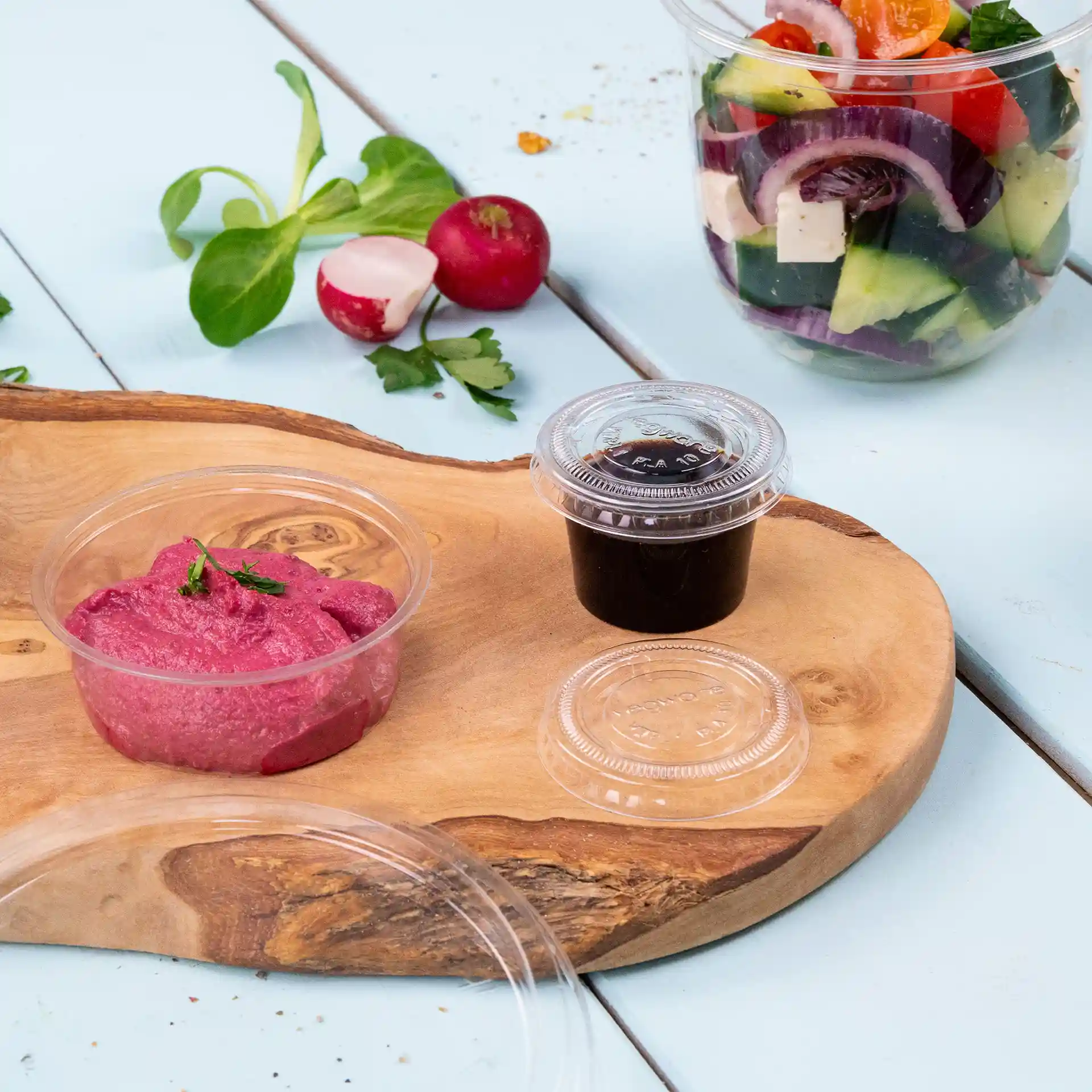 Afbeelding van een houten plank met een roze spread in een doorzichtige beker, een donkere saus in een andere doorzichtige beker en verse groenten op de achtergrond.