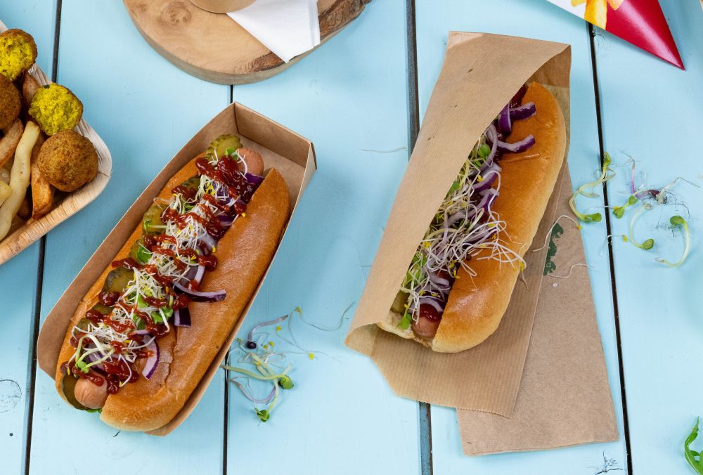 Twee hotdogs met verschillende toppings, waaronder augurken, uien en kiemen, geserveerd in papieren bakken op een blauwe tafel.