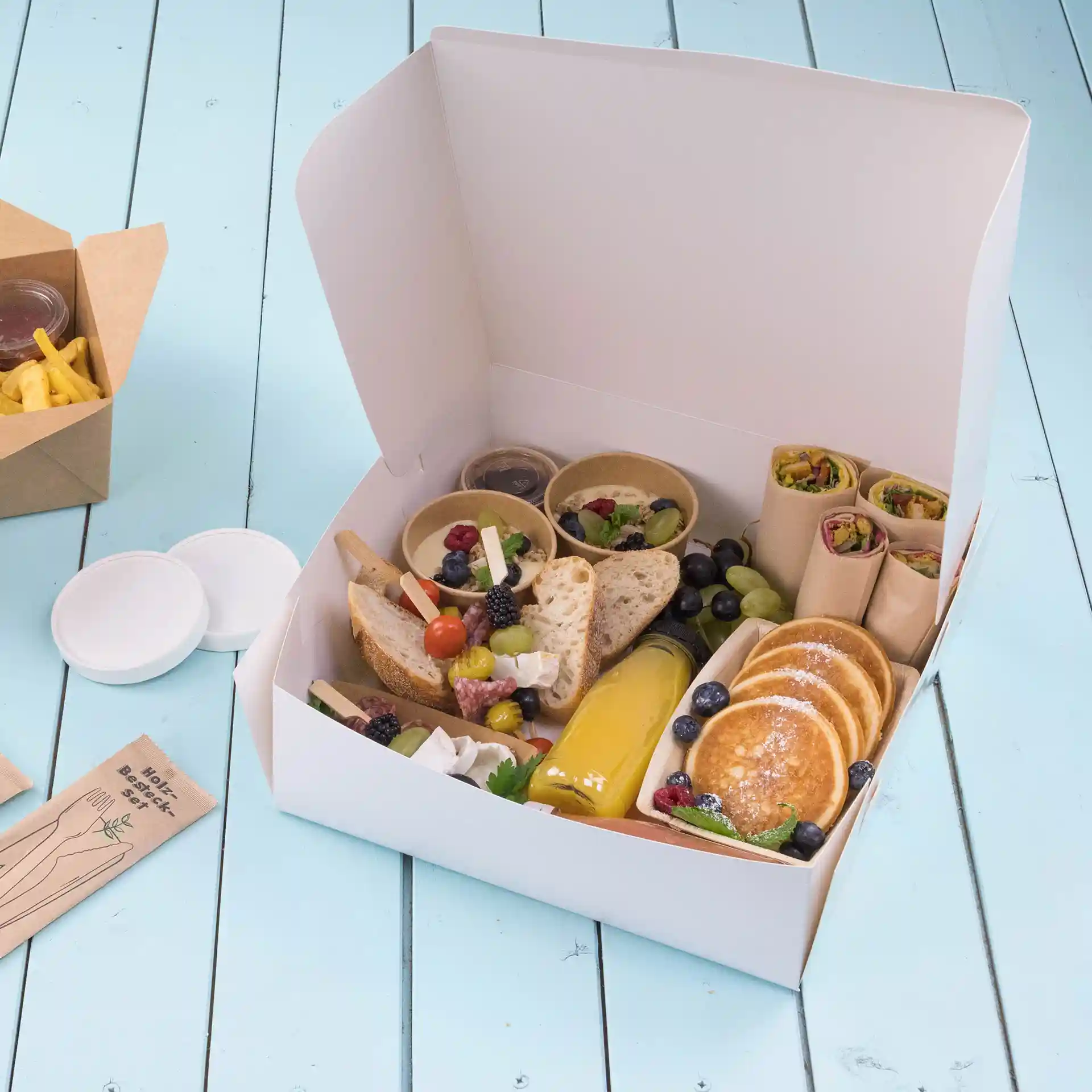 Witte cakebox gevuld met diverse voedingsmiddelen zoals brood, fruit, pannenkoeken en sauzen, op een blauwe houten ondergrond.