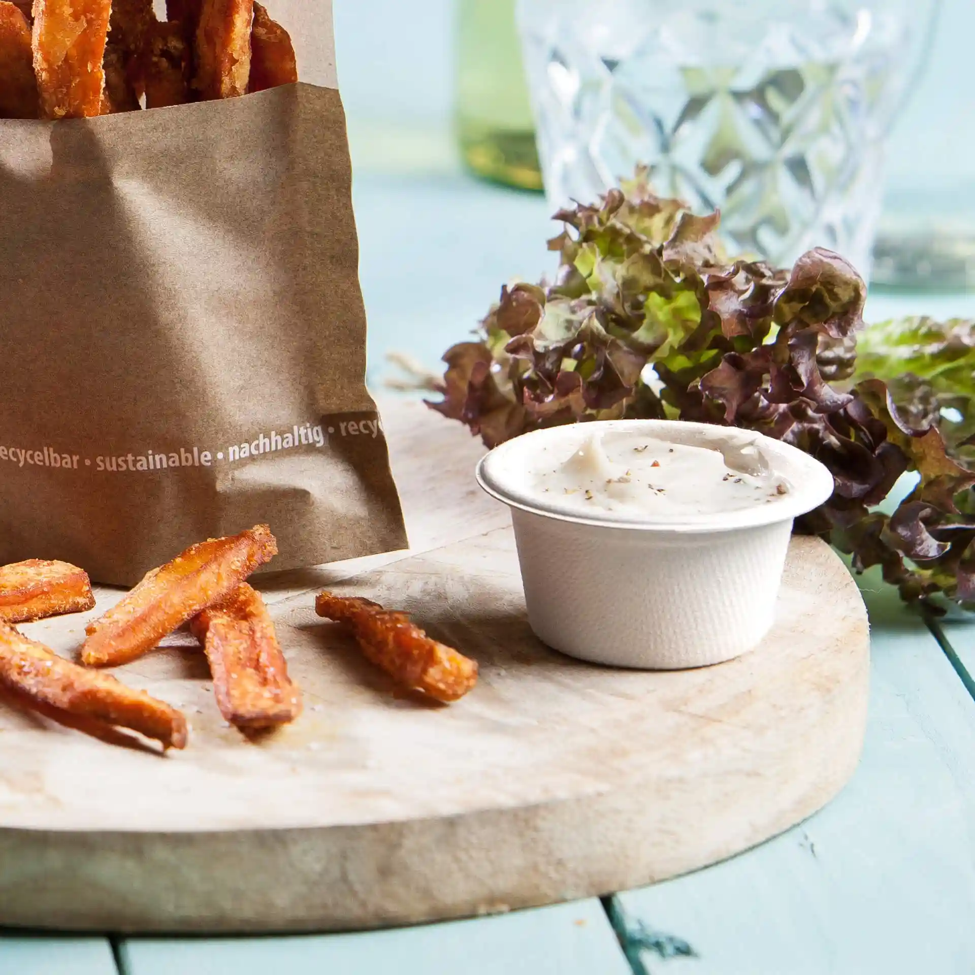 Afbeelding van een papieren zak met zoete aardappelfrites, een klein rond bakje dip en sla op een houten plank.
