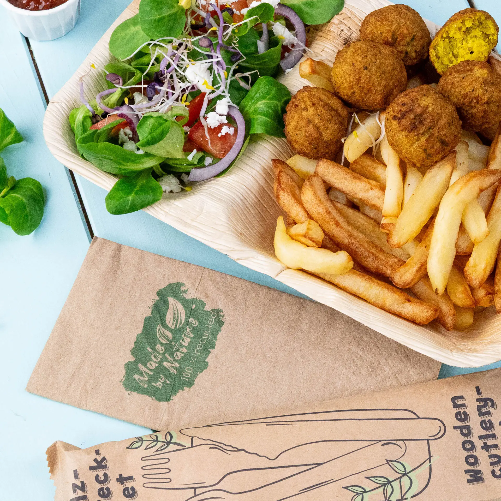 Een bord met falafel, friet en een gemengde salade; houten bestekverpakking zichtbaar; papieren servet met het label 'Made by Nature.'